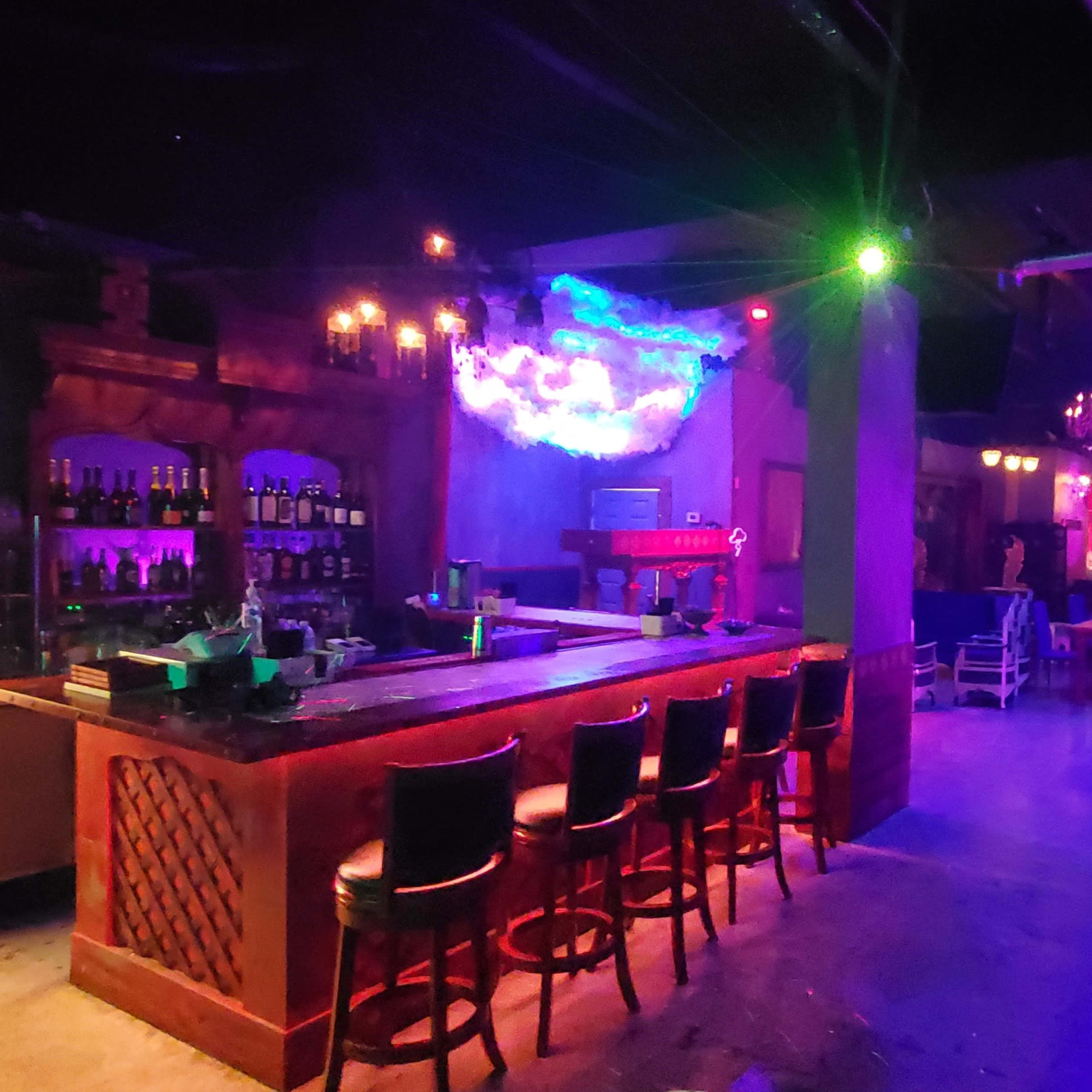 A dimly lit bar area with colorful lighting, featuring a long bar with stools and seating areas decorated with couches.