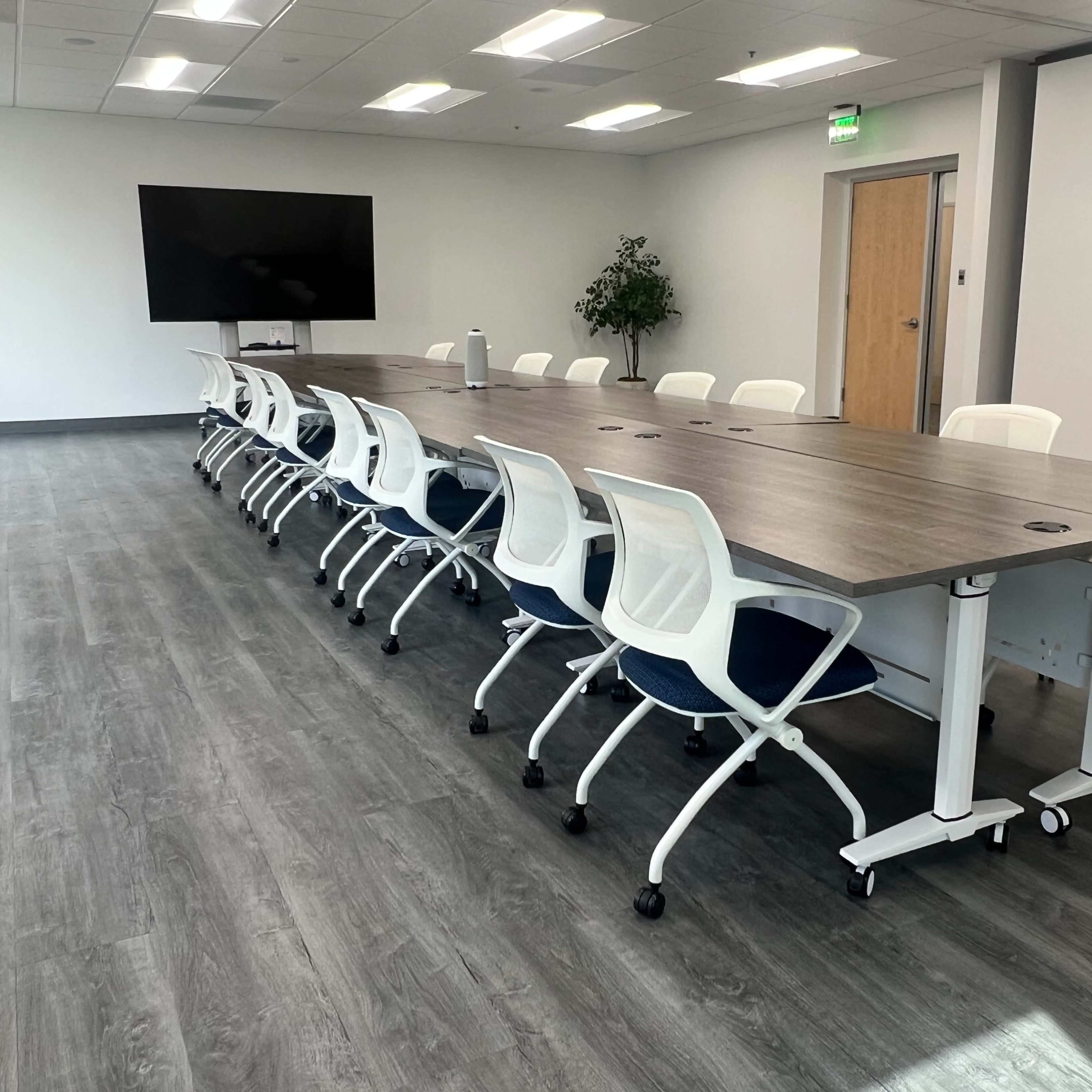 A long conference table with rolling white chairs is set up in a modern, brightly lit room that features a large screen and a potted plant.