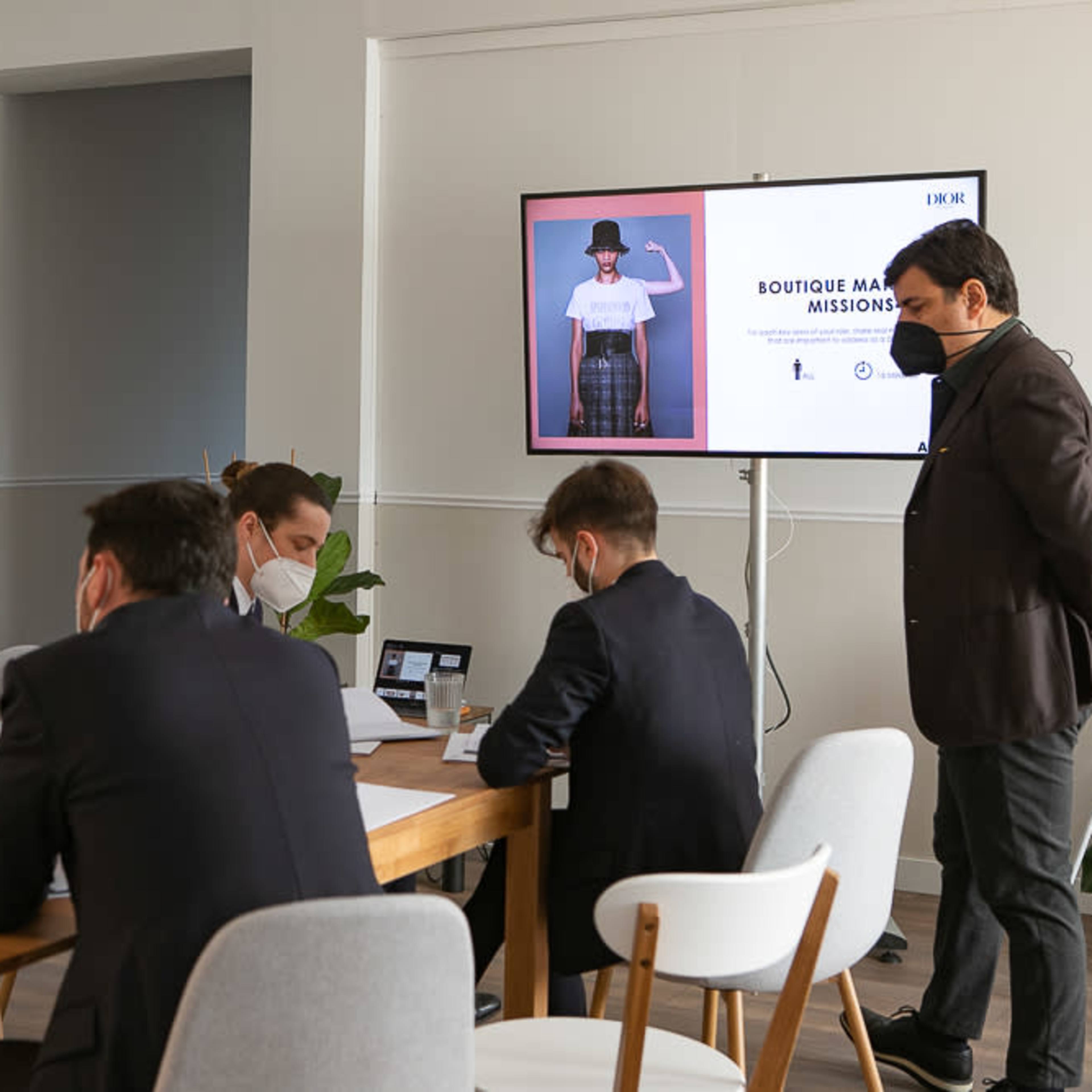 A group of five professionals in business attire is seated around a table, engaged in discussion, while a presentation is displayed on a screen.