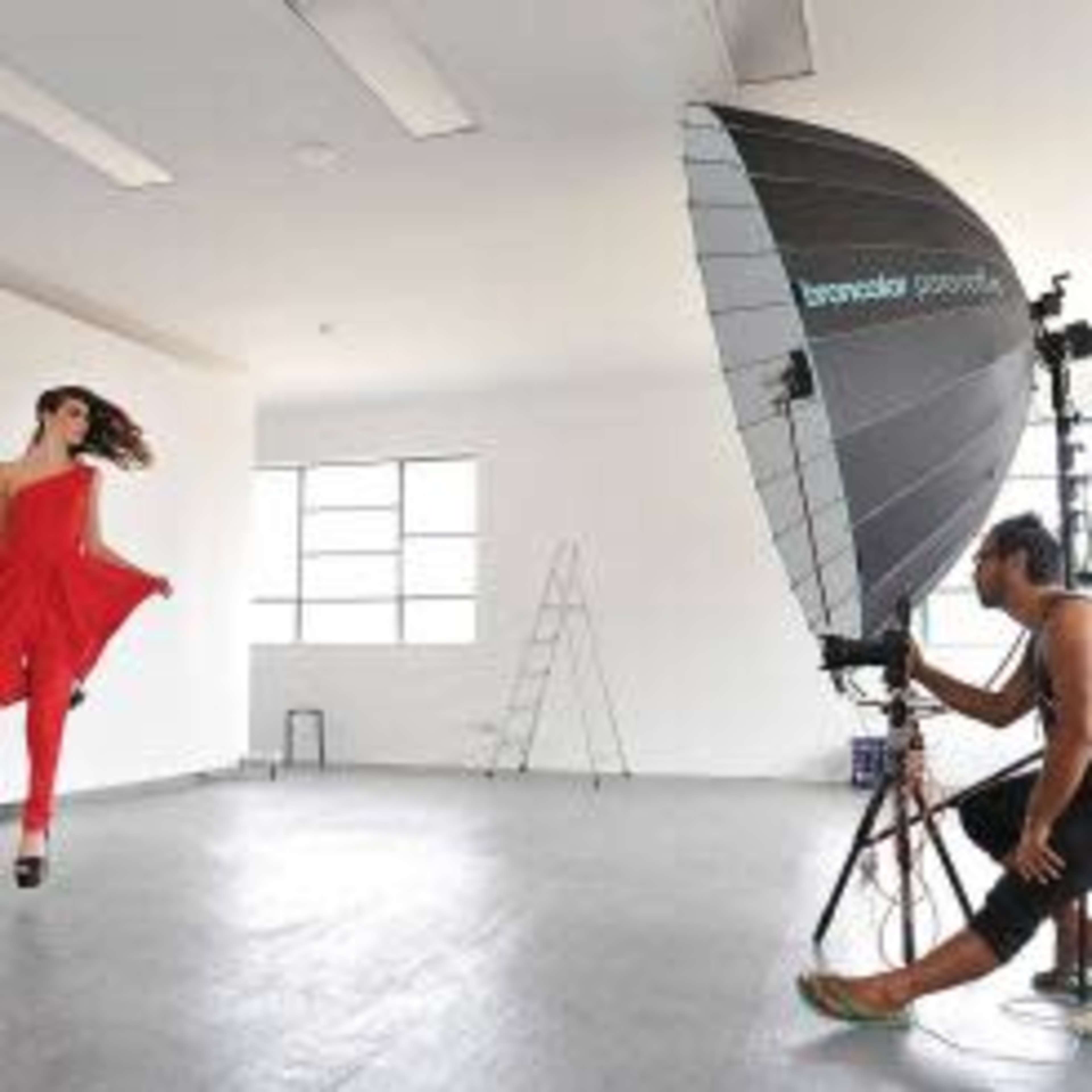 A model poses in a red outfit while a photographer adjusts lighting in a spacious studio.