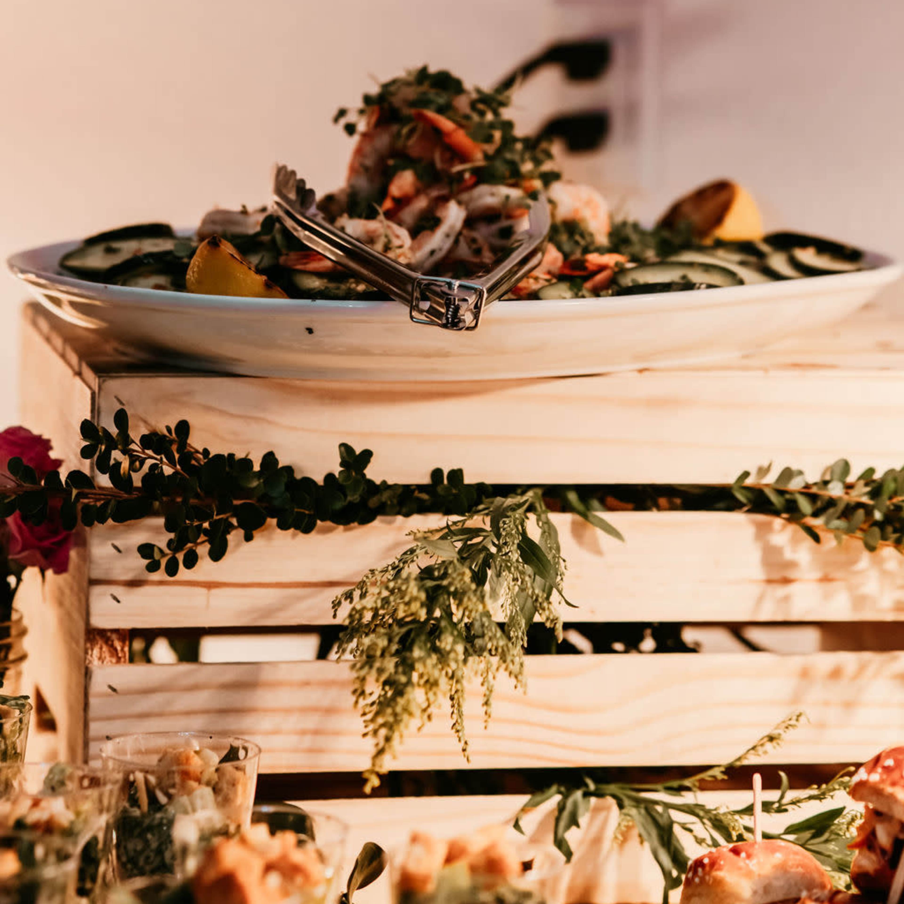 A large platter of seafood garnished with lemon slices is displayed on a wooden crate surrounded by greenery.