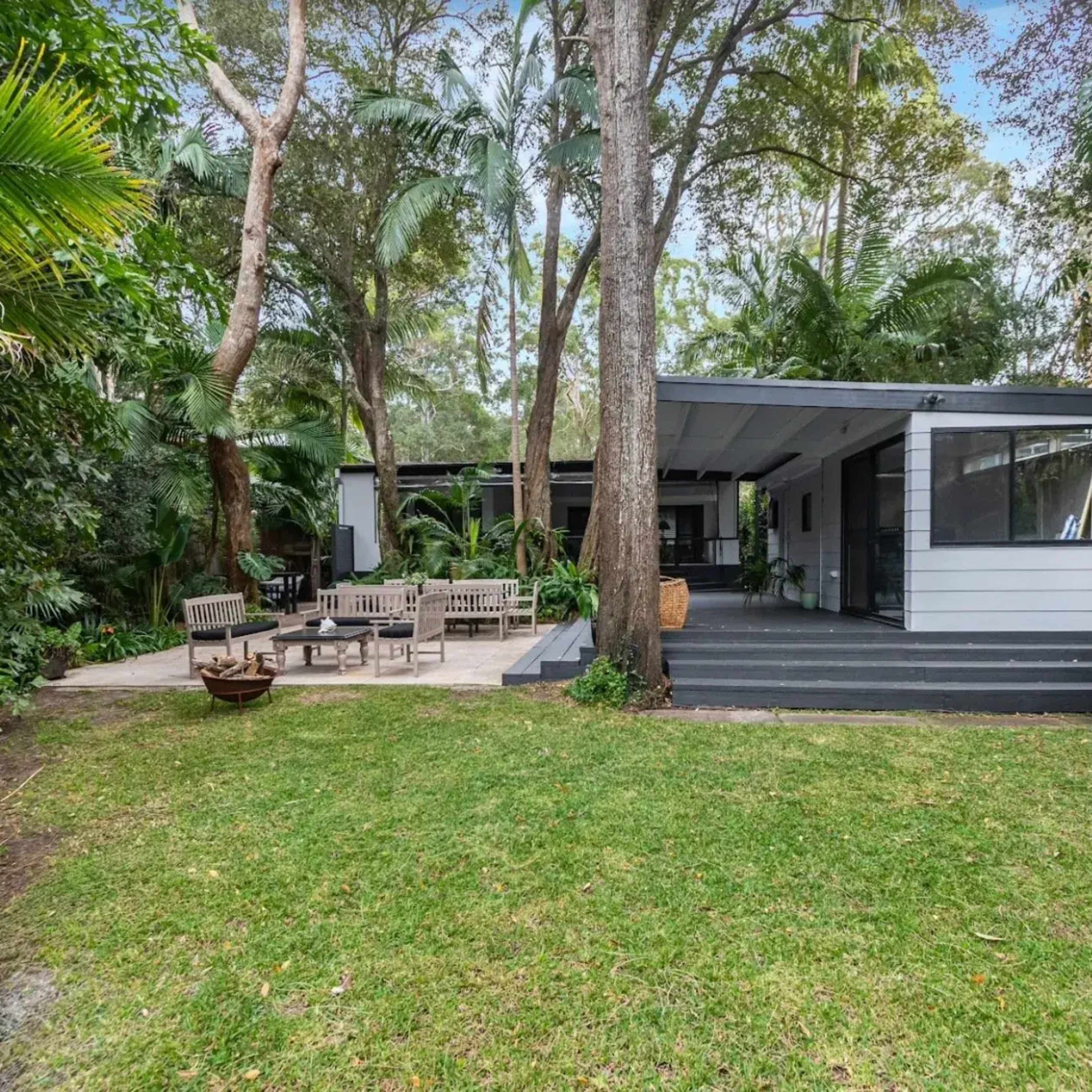 A modern house with a gray exterior sits surrounded by lush greenery and a patio area with outdoor seating.