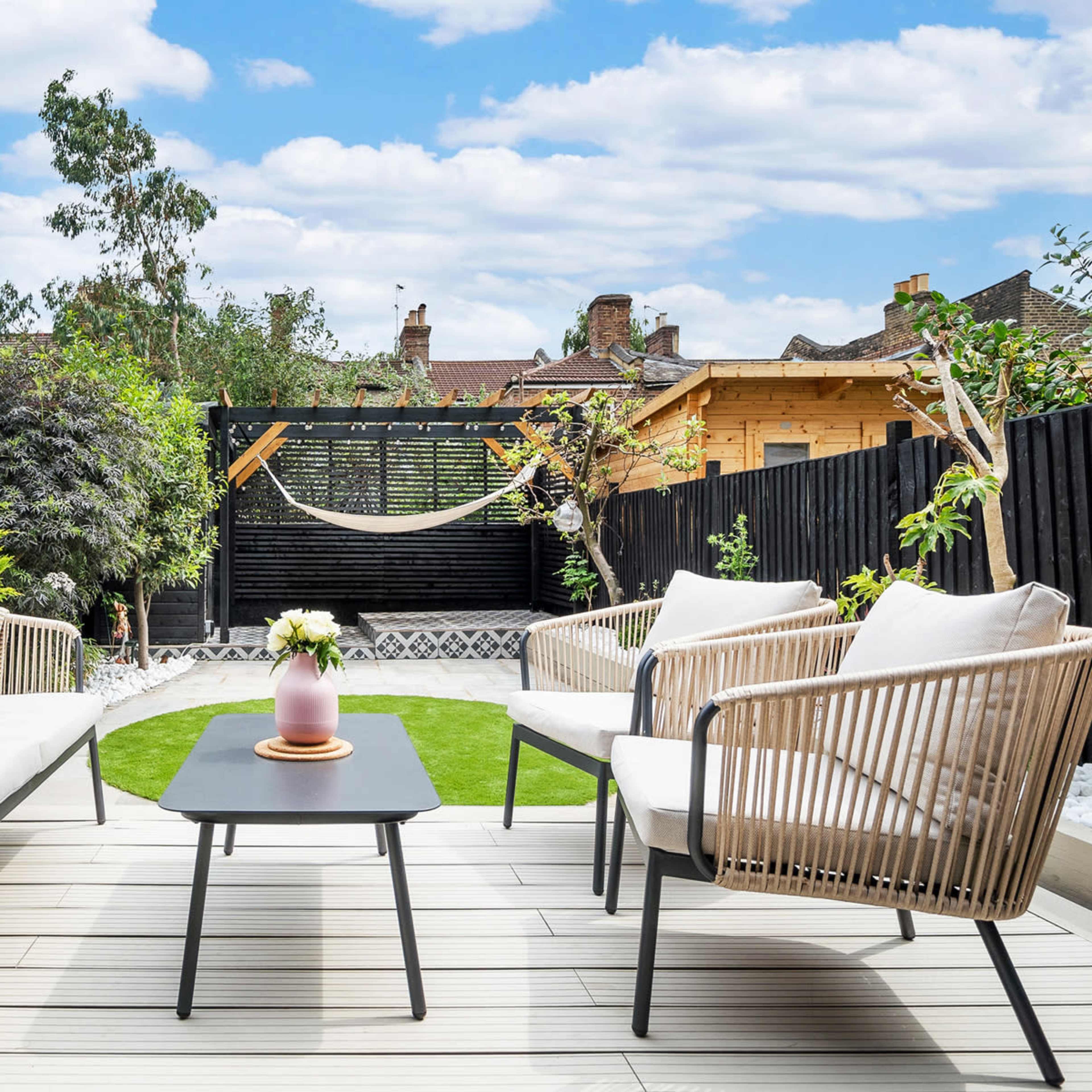 A landscaped garden area with two modern seating arrangements and a small table, featuring a hammock in the background and greenery along the perimeter.