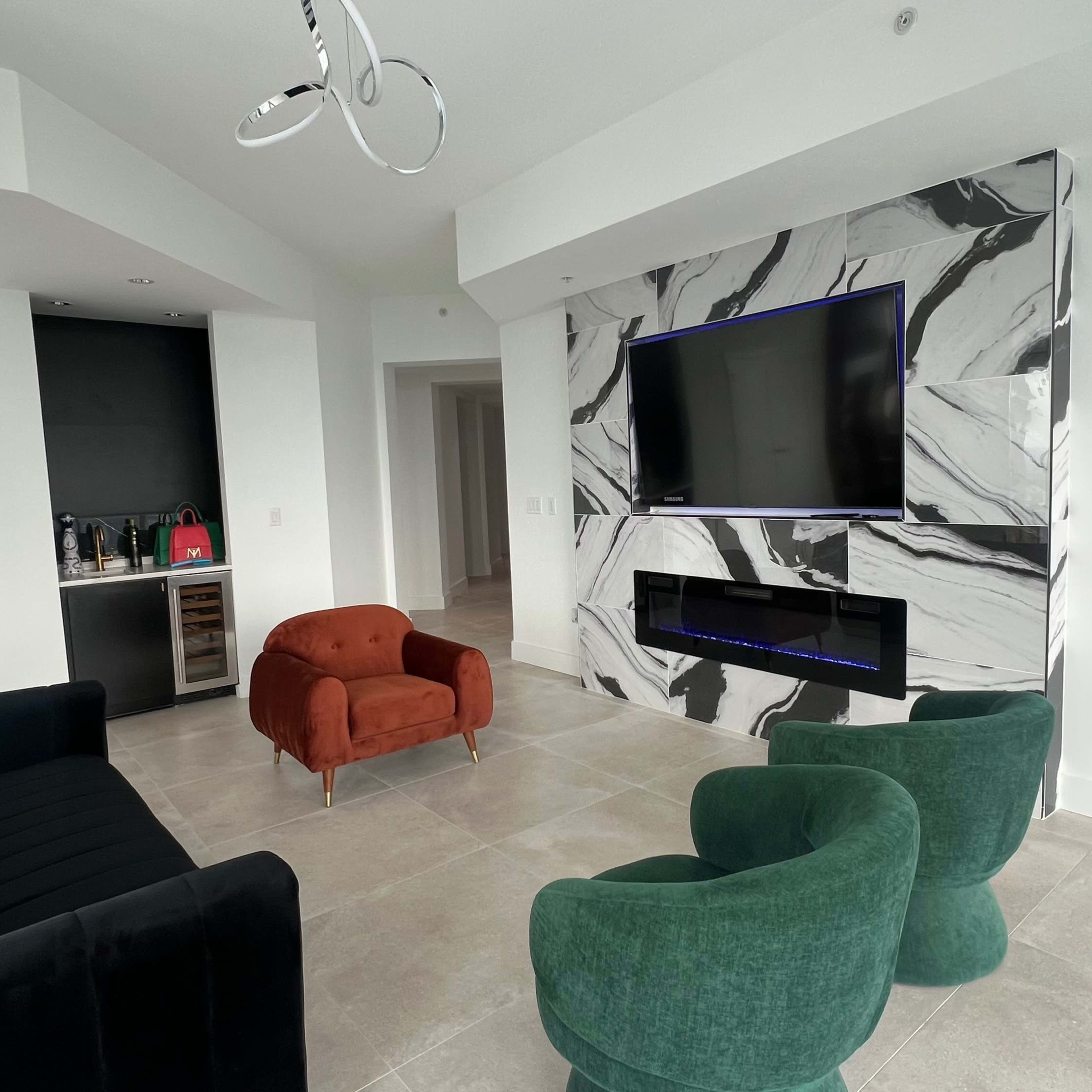 A modern living space with a marble-featured wall, a wall-mounted television, two green chairs, an orange armchair, and a black sofa.