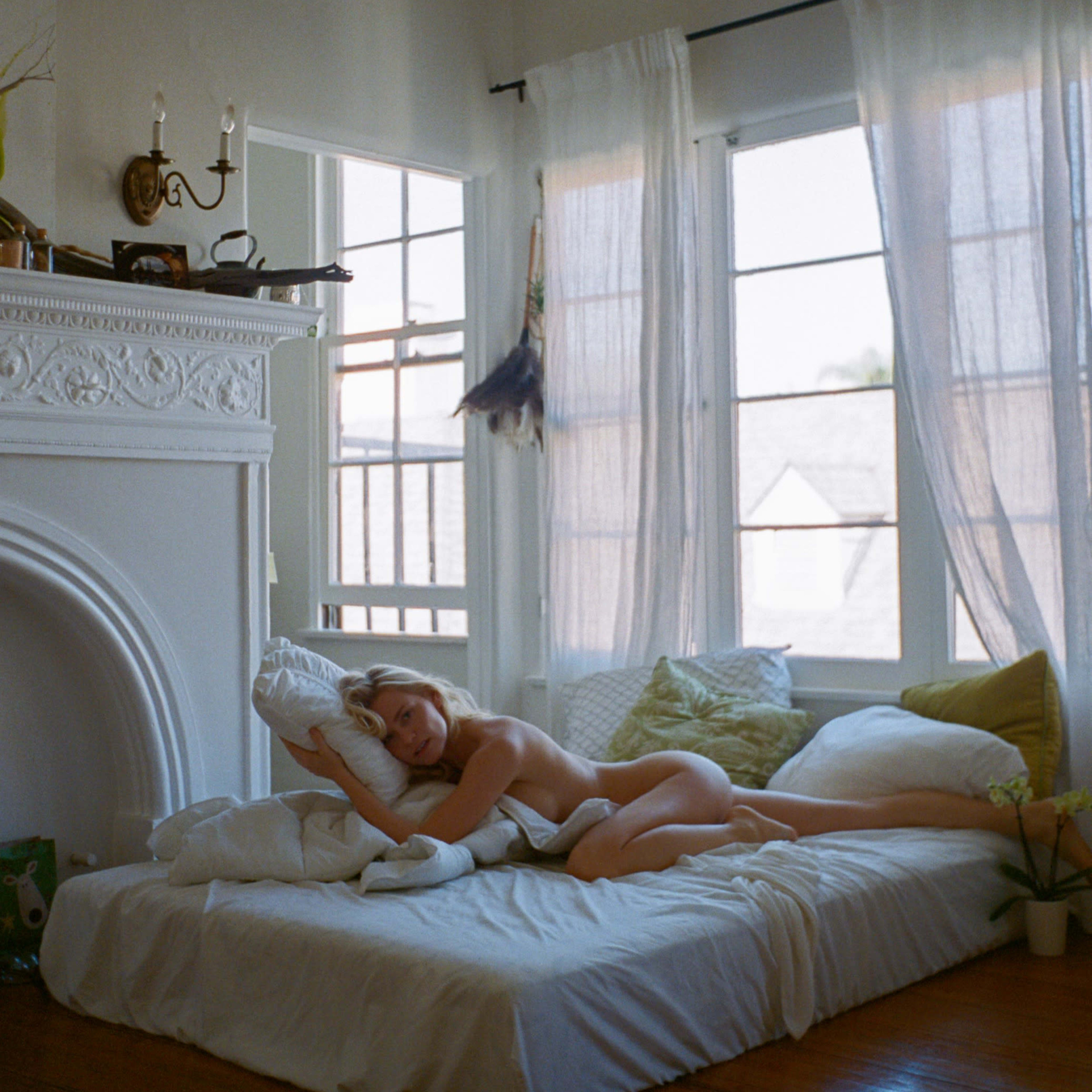 A person is lying on a bed in a well-lit room with large windows and light-colored curtains.