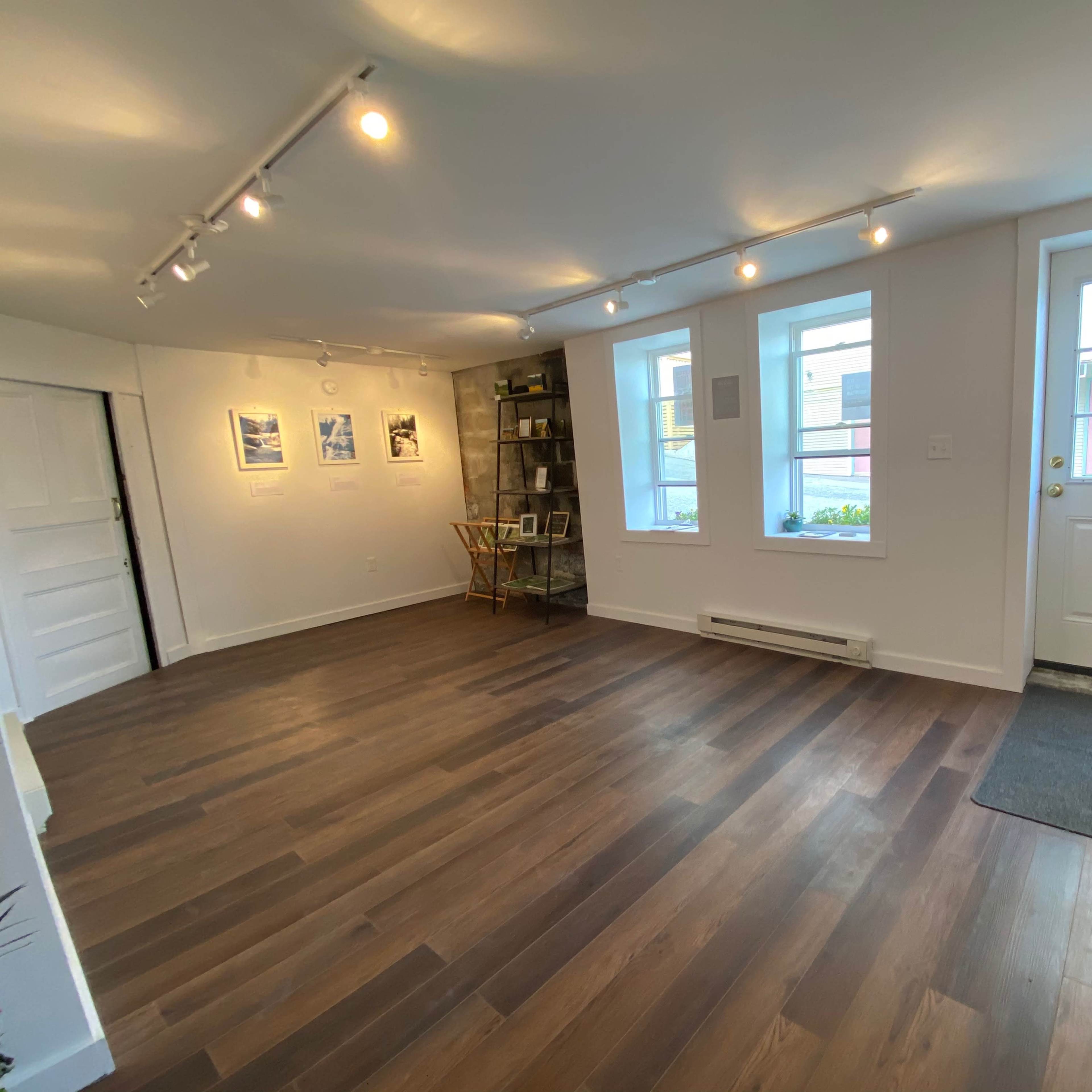 A bright, minimally furnished room with wooden flooring, three windows, and a door leading outside.