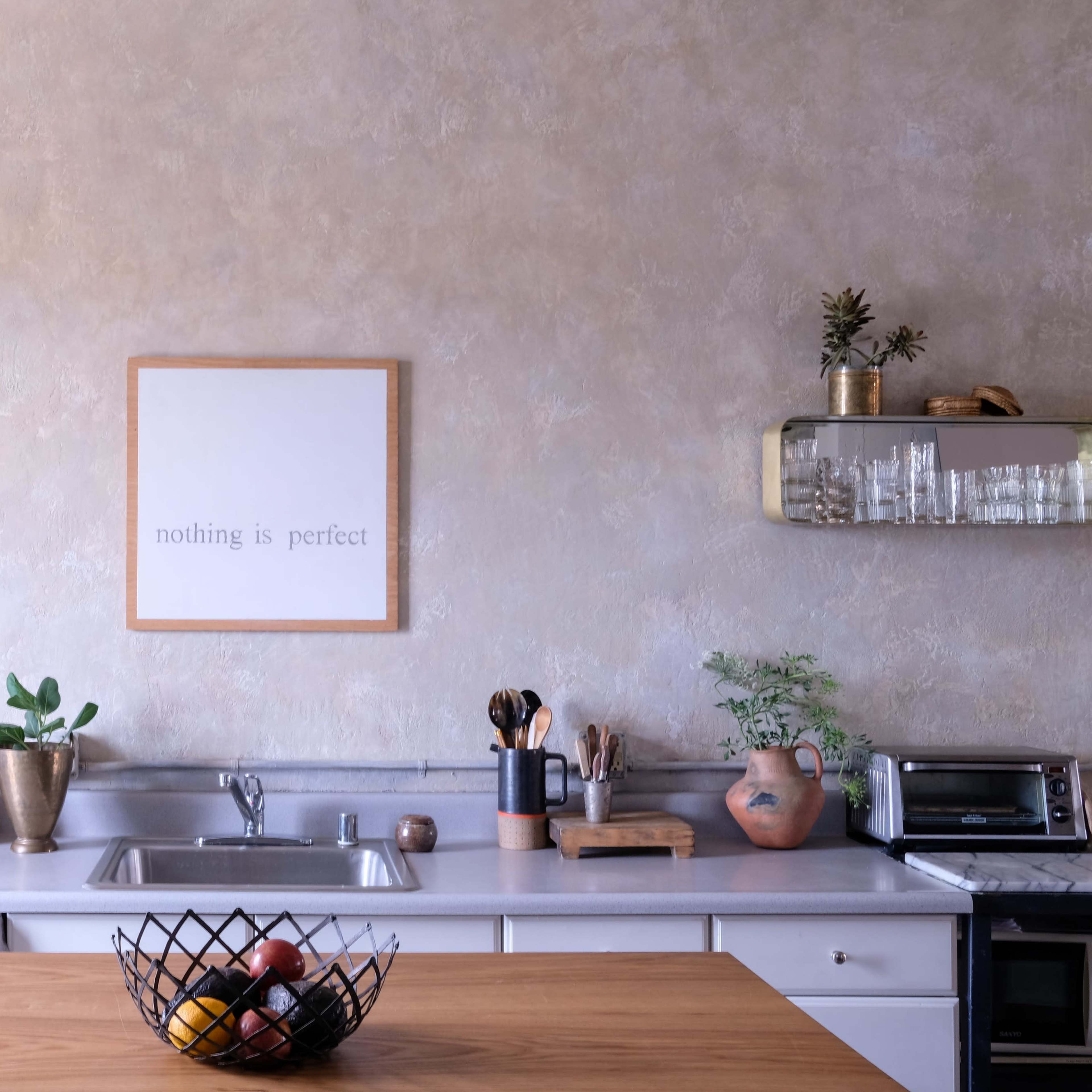 A modern kitchen features a wooden table, a sink with utensils, and a decorative wall with a framed quote.