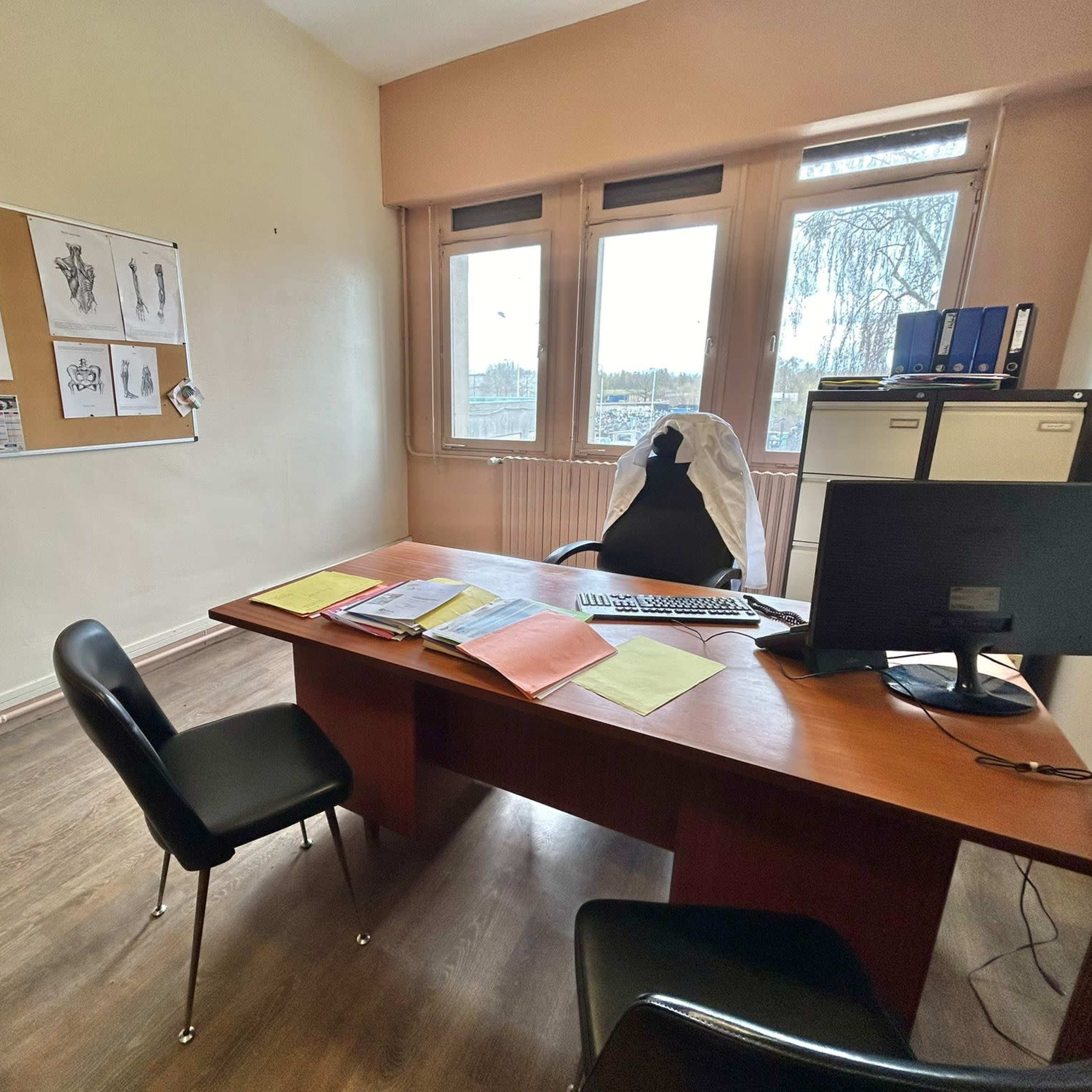 A spacious office features a large wooden desk with a computer and various documents, alongside a chair, two black chairs, and windows that provide natural light.