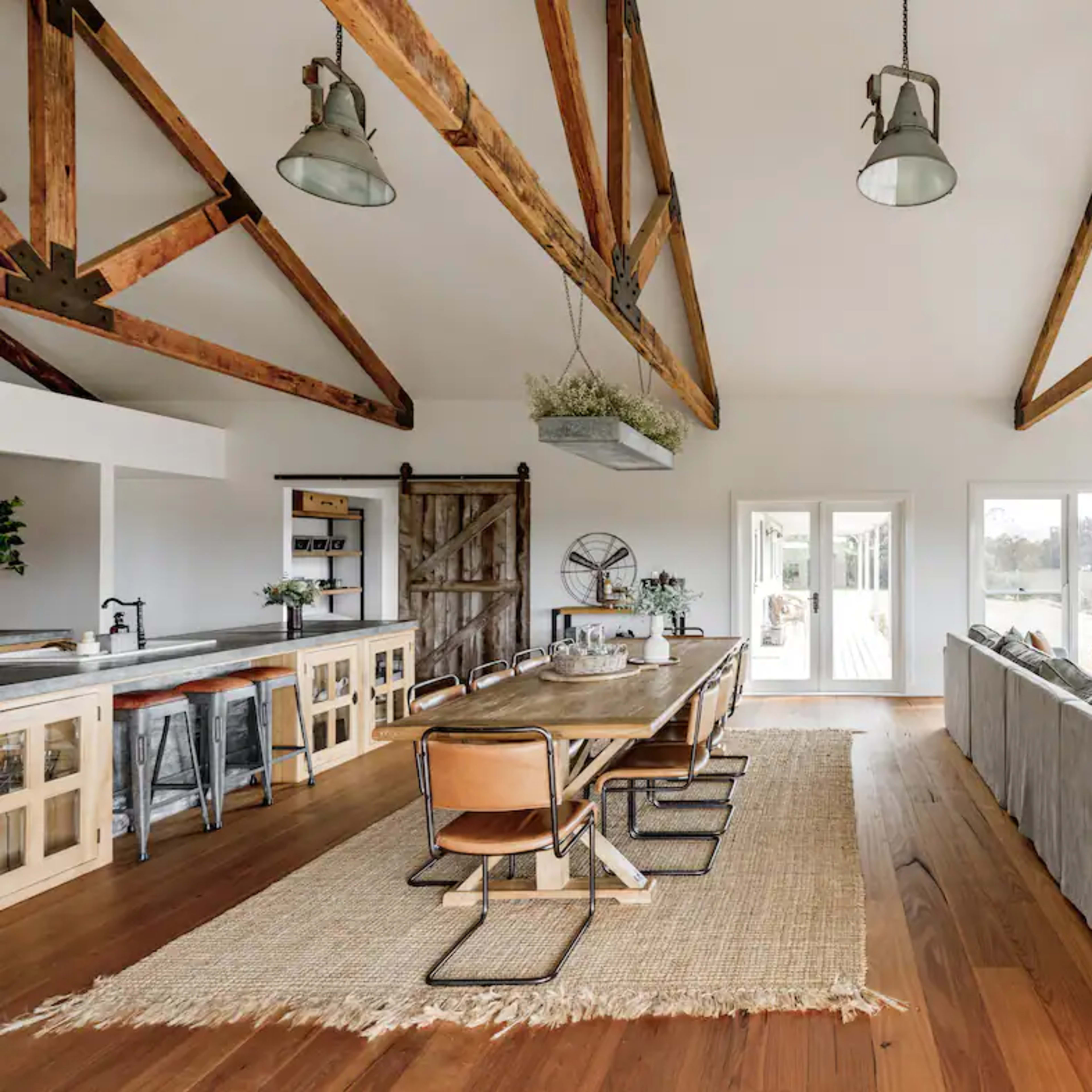 A spacious living area featuring exposed wooden beams, a large dining table, and large windows overlooking a natural landscape.