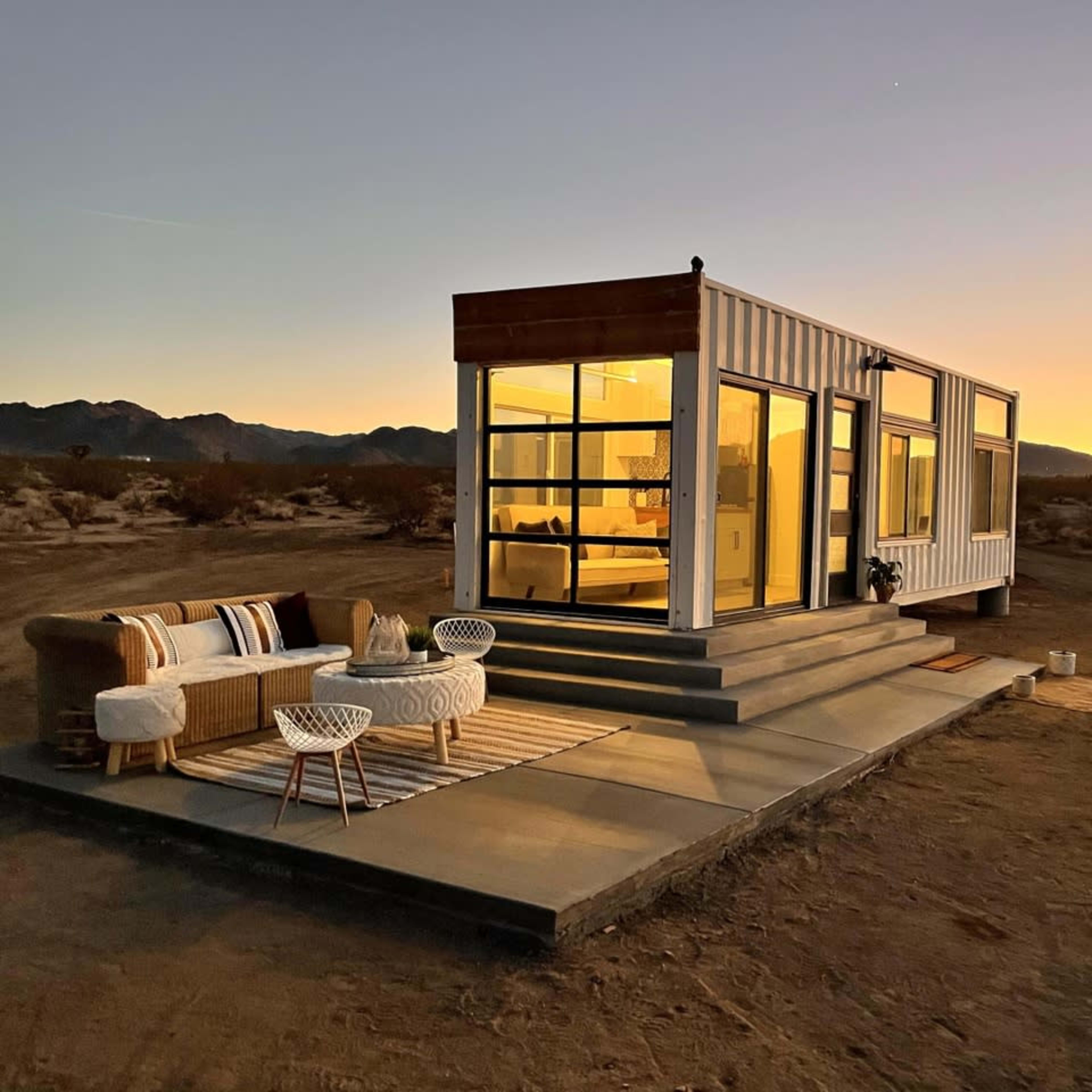 A modern, container-style home with large windows is situated on a desert landscape at sunset, accompanied by outdoor seating.