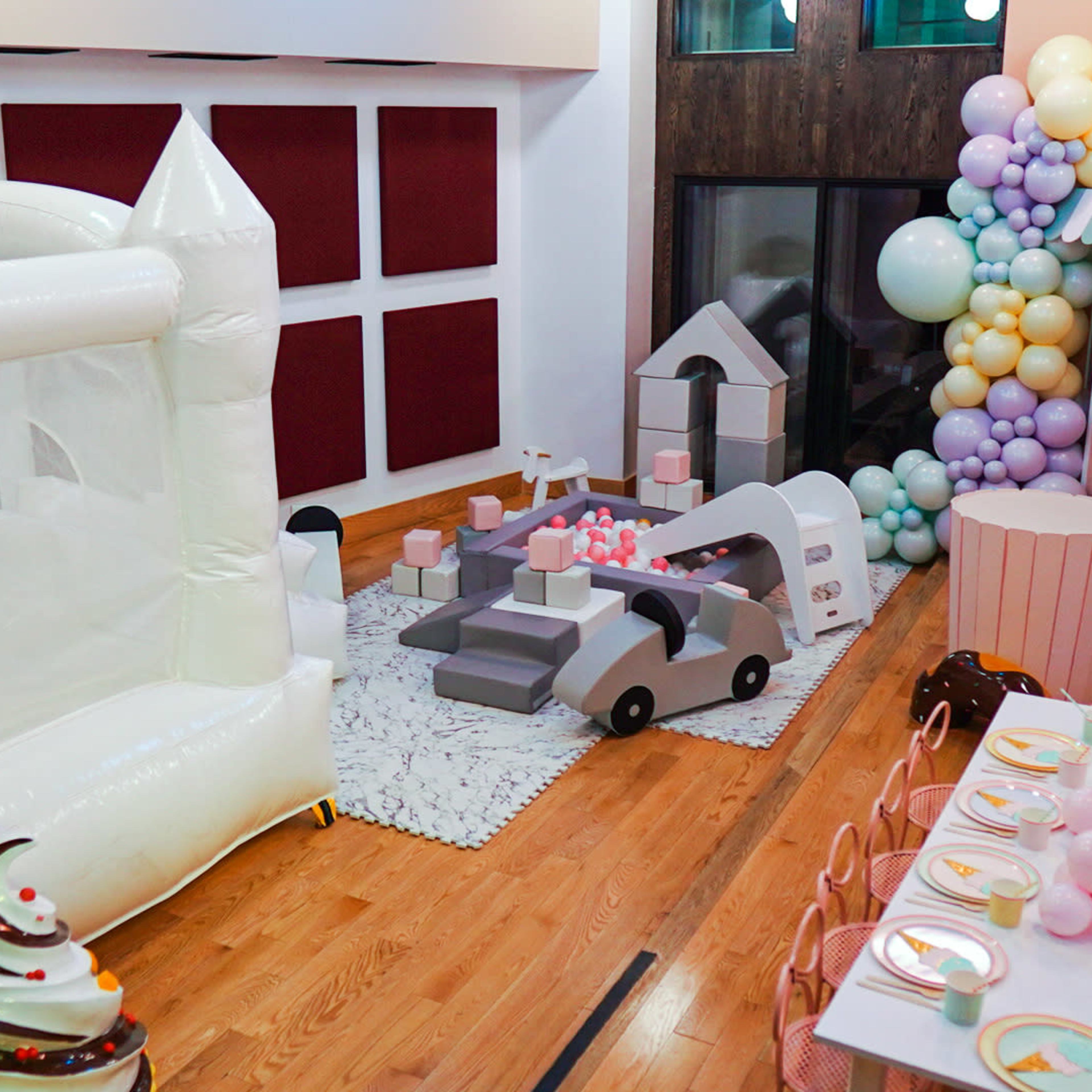 A children's party setup featuring an inflatable bounce house, a play area with toy cars and a miniature house, and a decorated table with pastel-colored plates and a balloon arch.