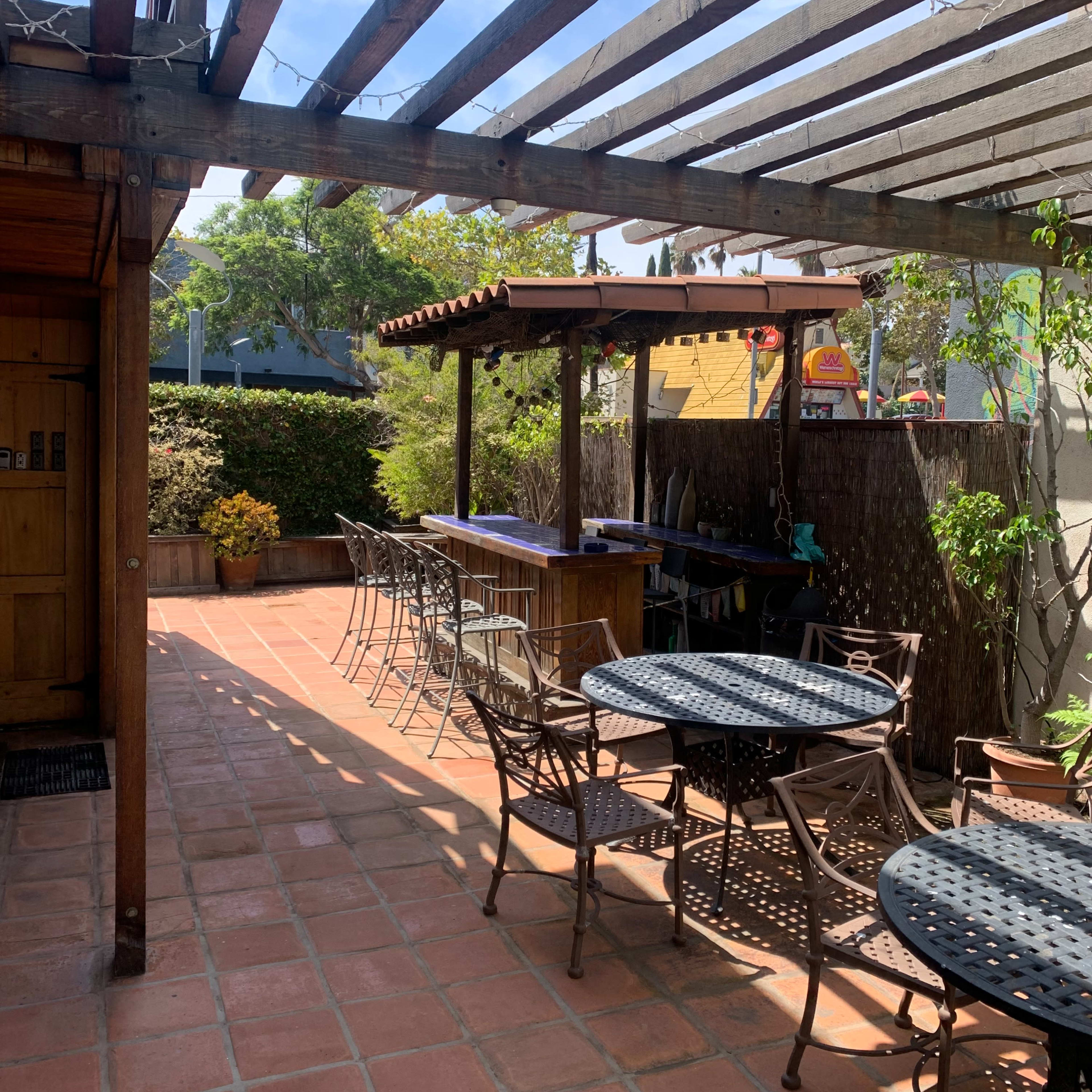 A shaded outdoor patio features wooden pergolas, metal tables and chairs, and potted plants on a terracotta tiled floor.