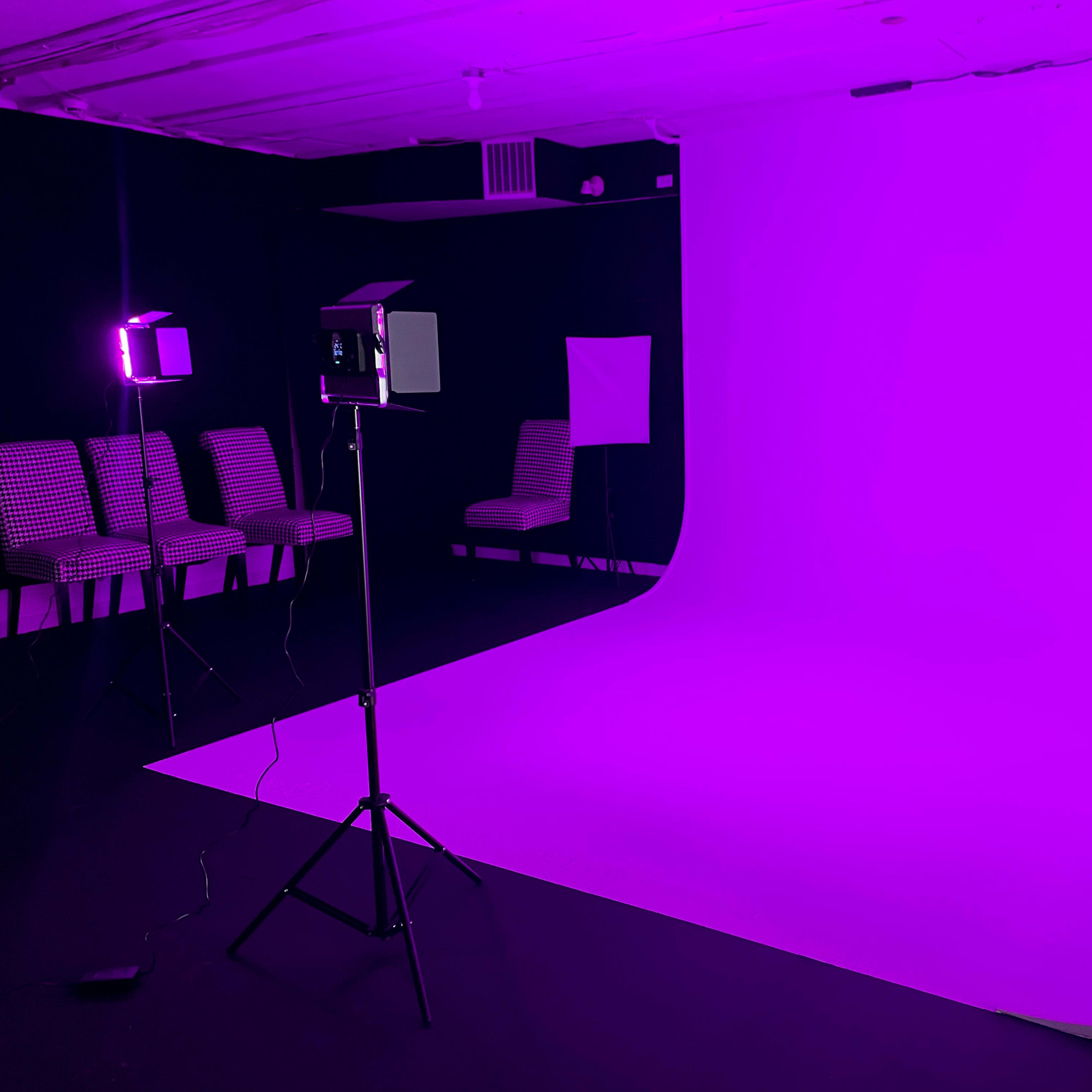 A photography studio with a purple backdrop, a light stand, and several empty chairs arranged against a dark wall.