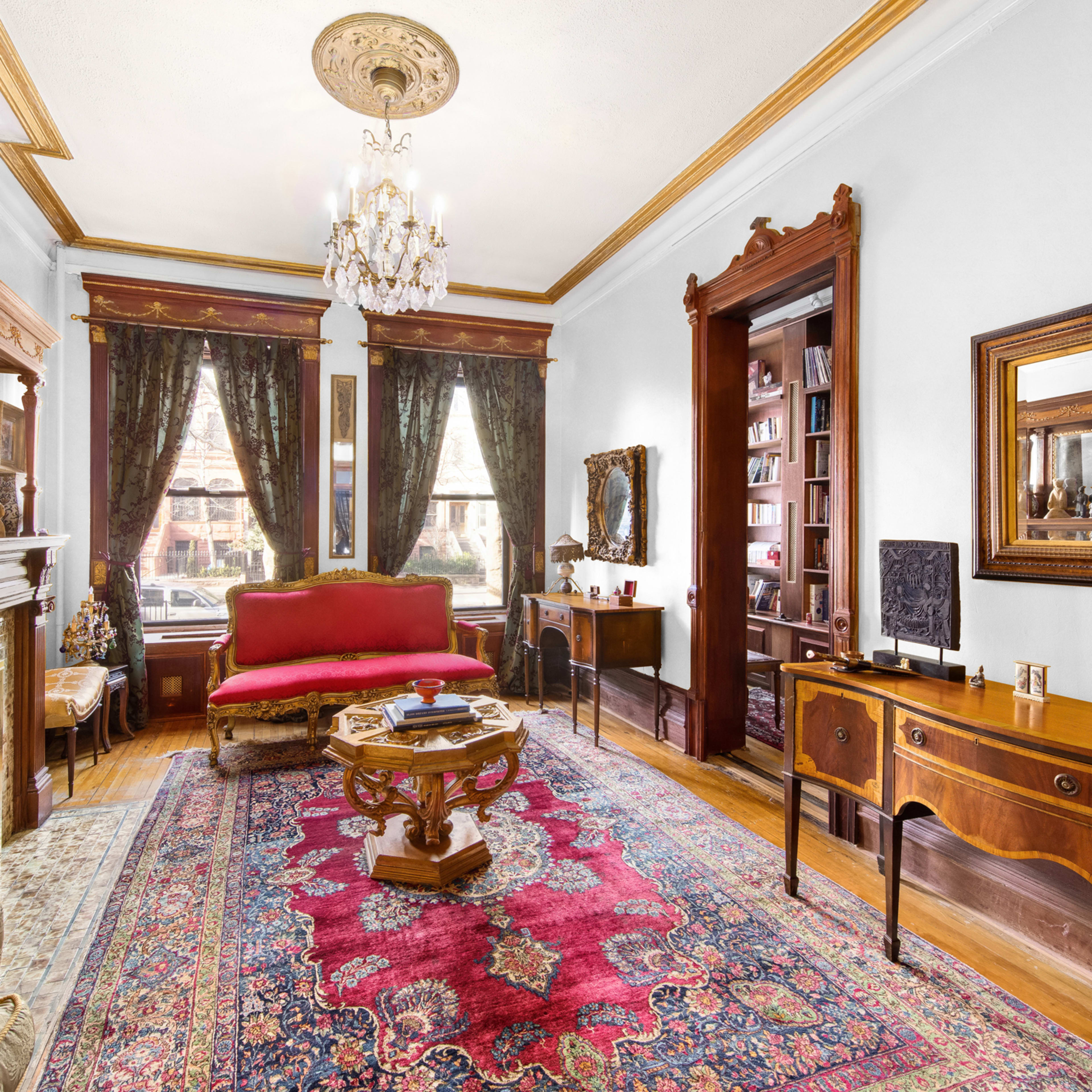A vintage living room featuring ornate wood furniture, a patterned rug, a chandelier, and large windows with dark curtains.