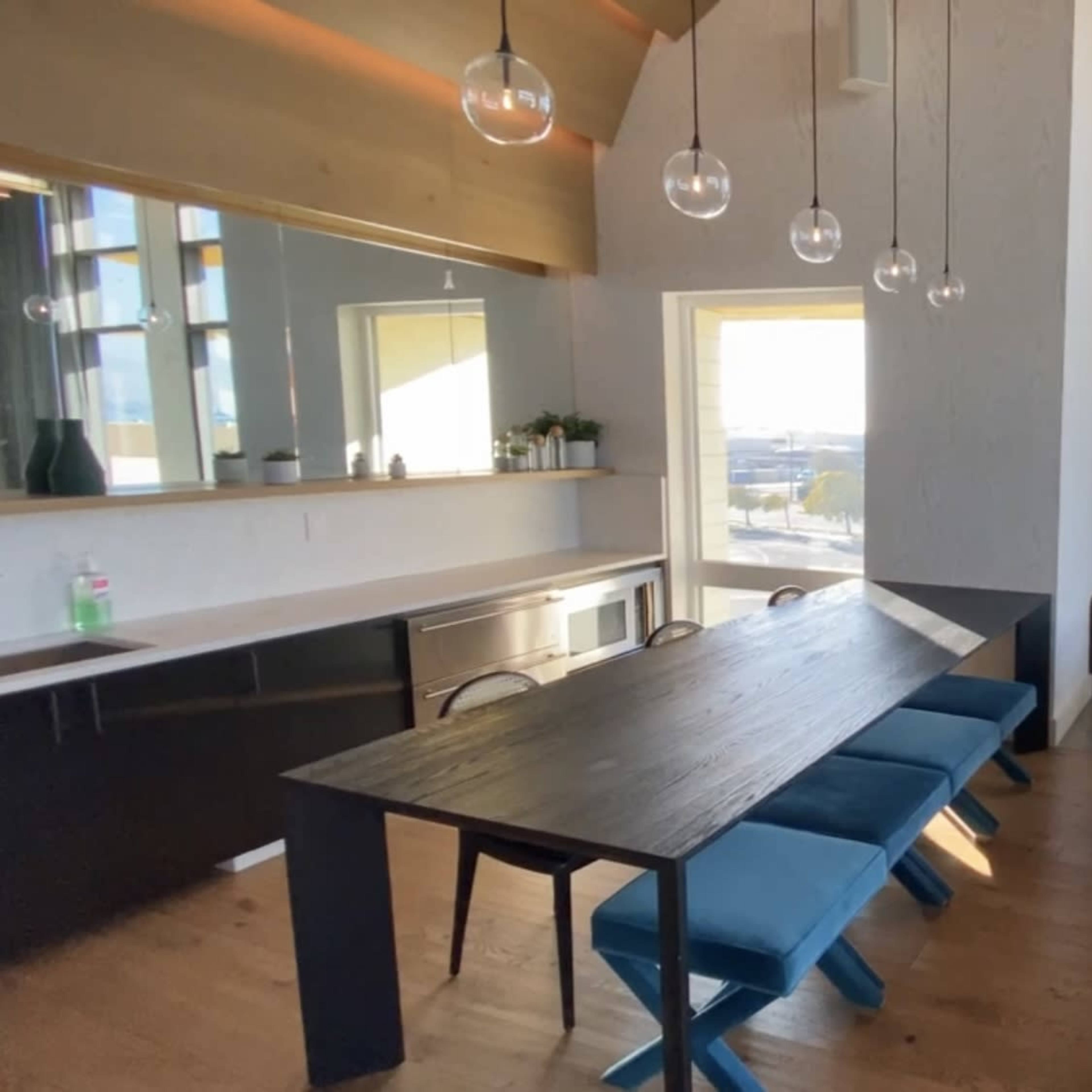 A modern living space featuring a dining table with six blue chairs, a dark sofa, large windows with yellow shades, and a kitchen area in the background.