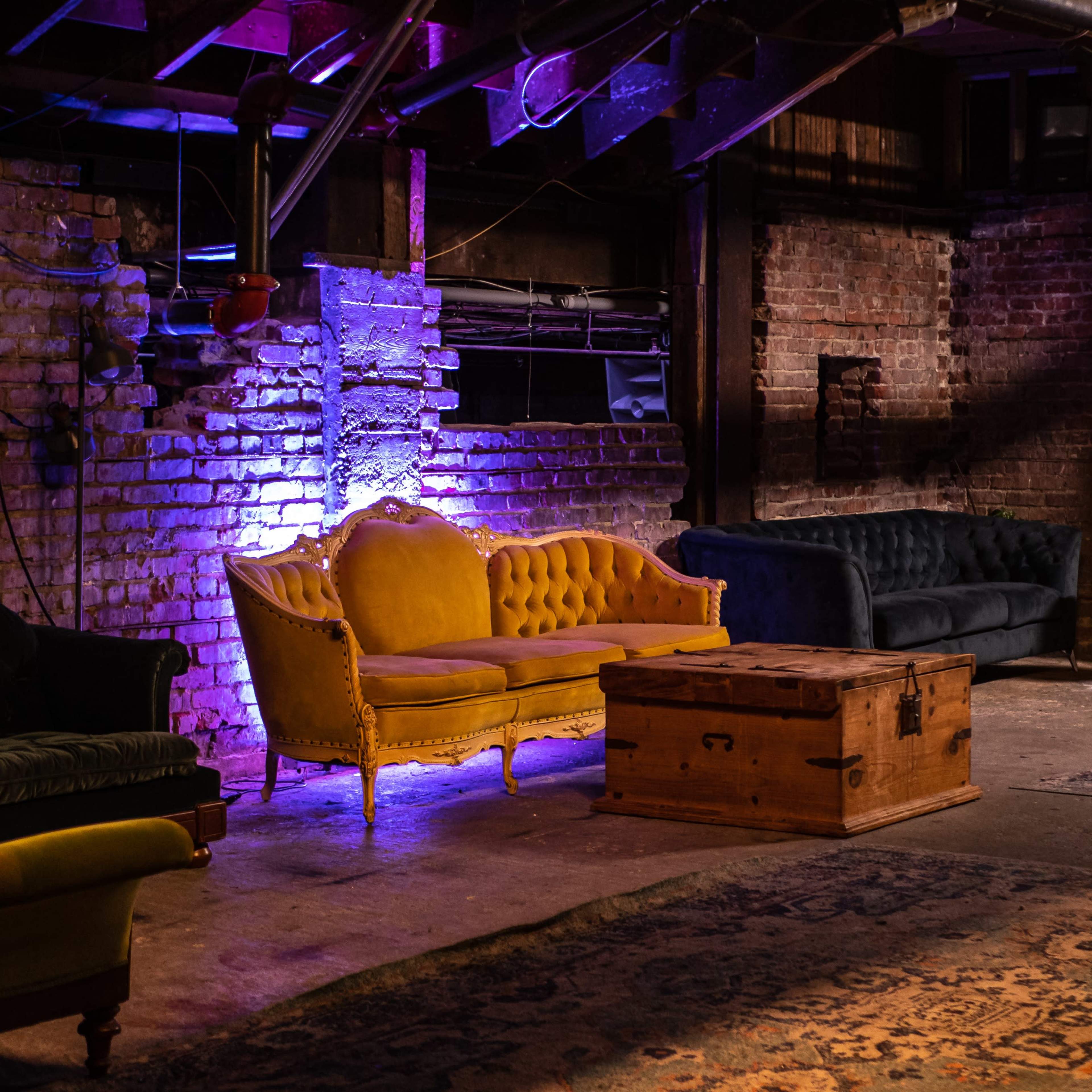 A dimly lit indoor space with various vintage-style sofas, a wooden coffee table, and area rugs against a backdrop of exposed brick walls.