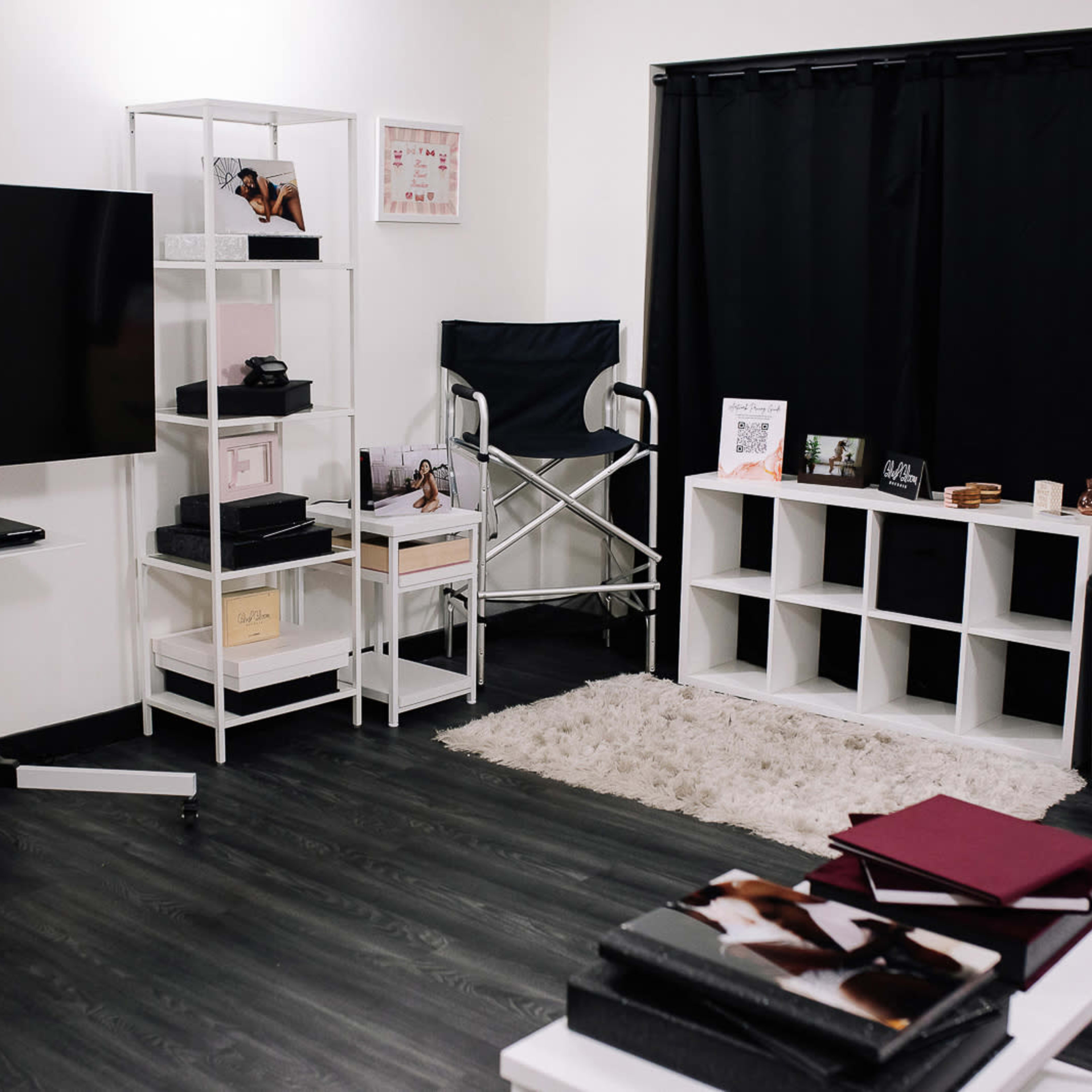 A modern, minimalist room featuring a large television on a stand, a white shelving unit with various items, a director's chair, and a stylish black curtain.