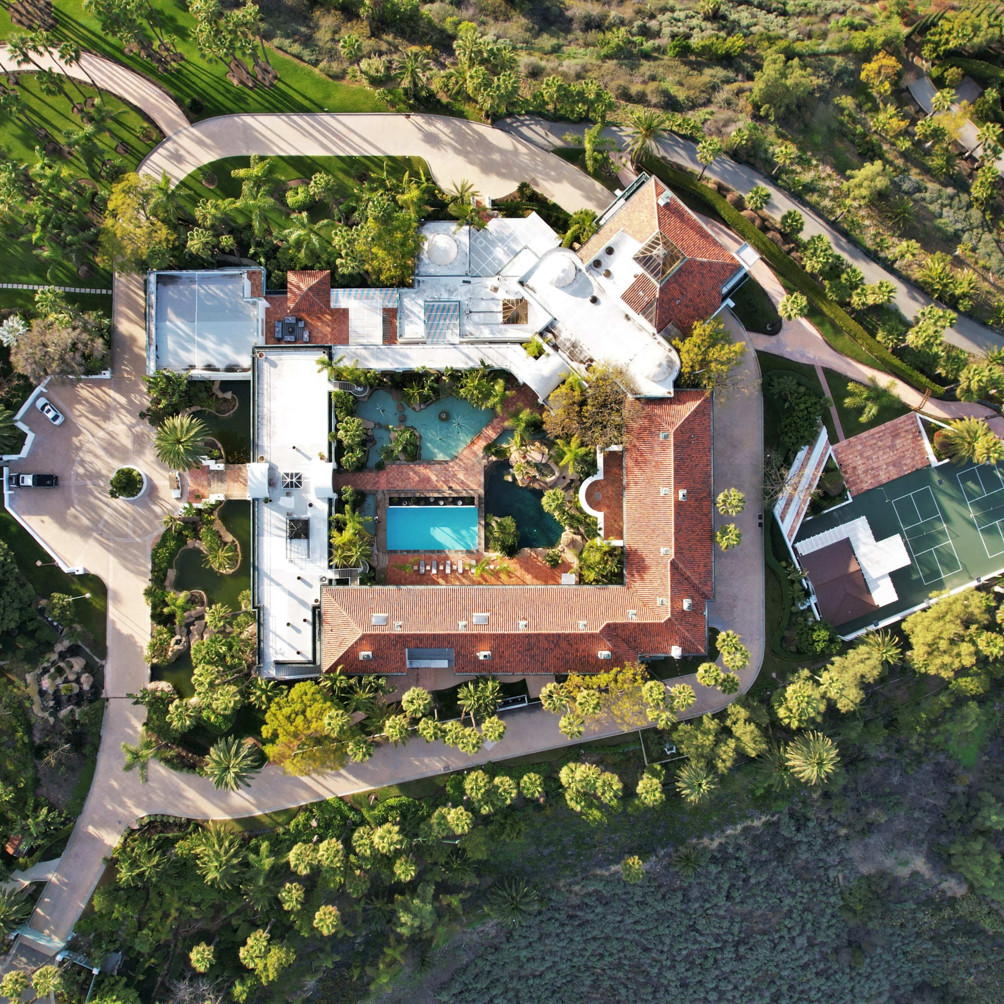 An aerial view of a sprawling estate features a main house with a pool, tennis court, and landscaped gardens surrounded by trees.