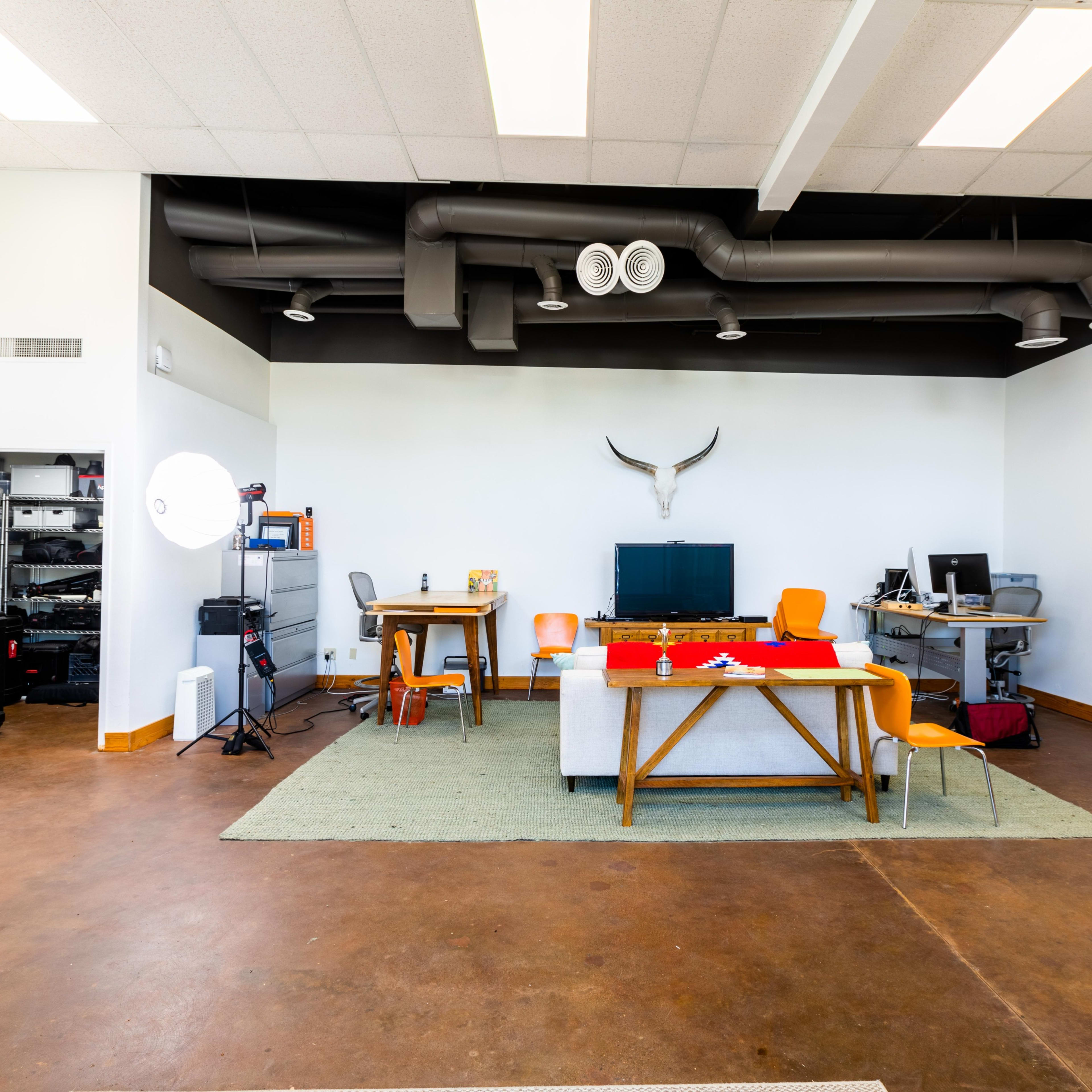 An open office space featuring two wooden tables, orange chairs, and a wall-mounted animal skull, with a large window and exposed ductwork on the ceiling.