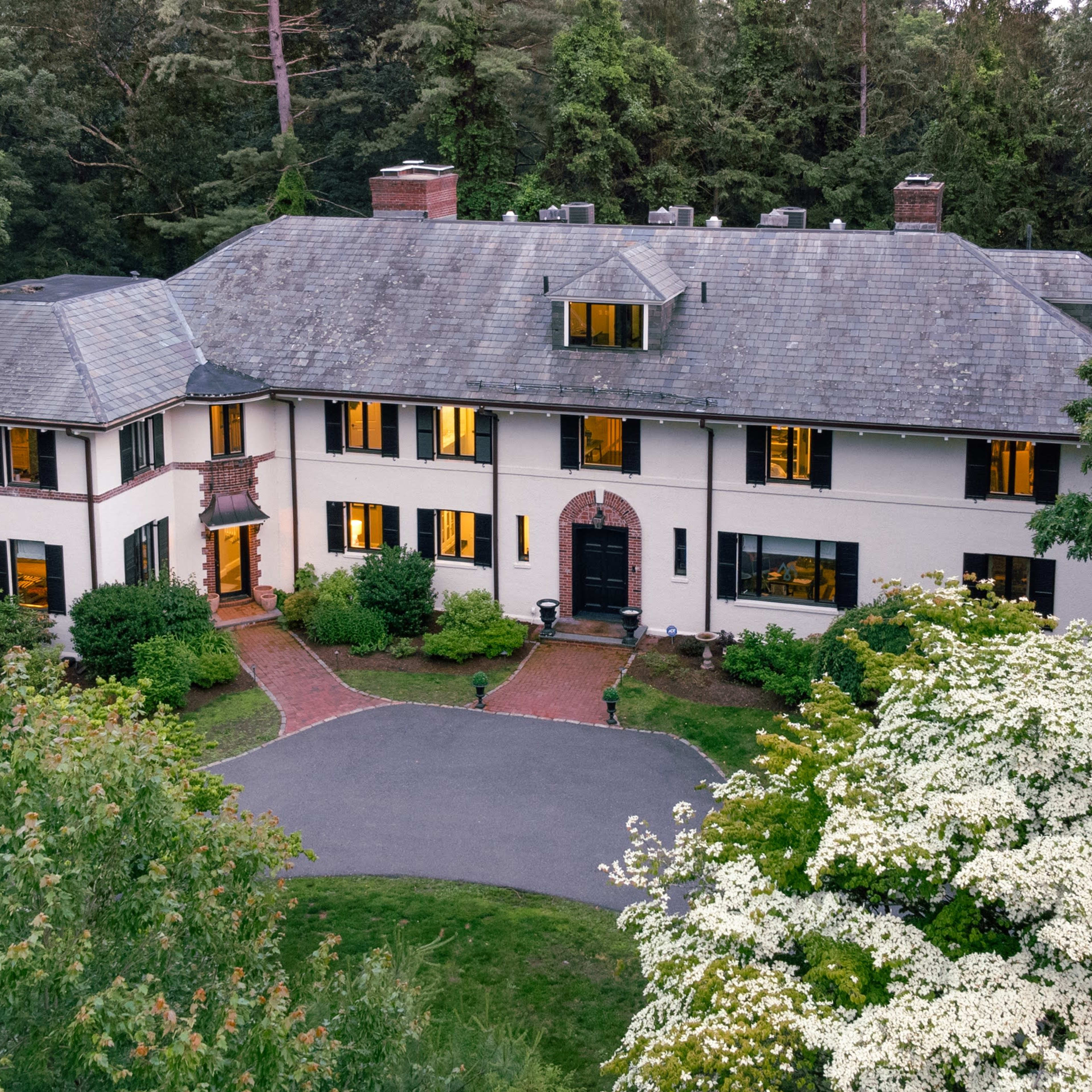 A large, multi-story house surrounded by trees and landscaping, with a winding driveway and large windows illuminated in the evening light.