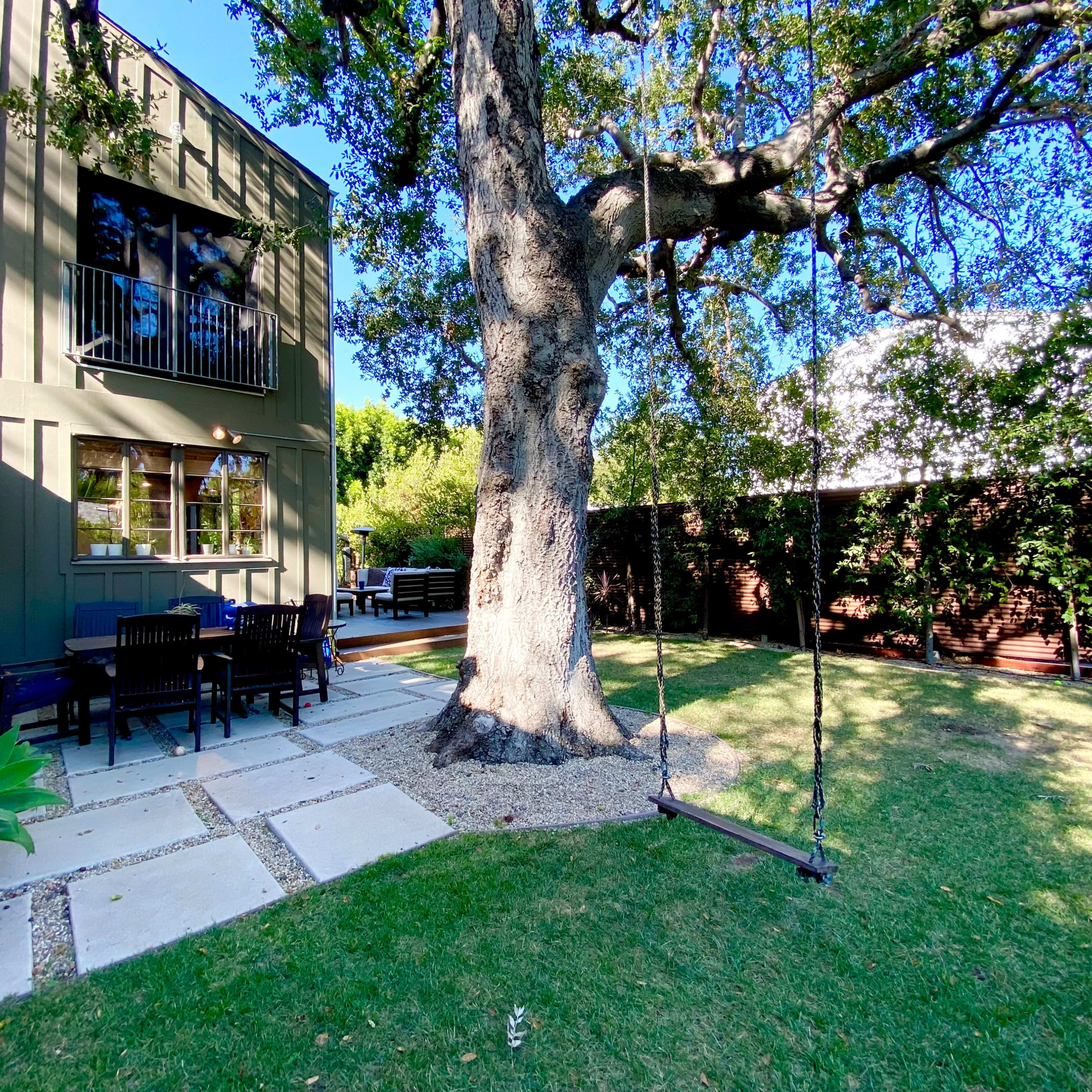 A large tree with a swing stands in a backyard adjacent to a house, featuring a stone pathway and a patio set.