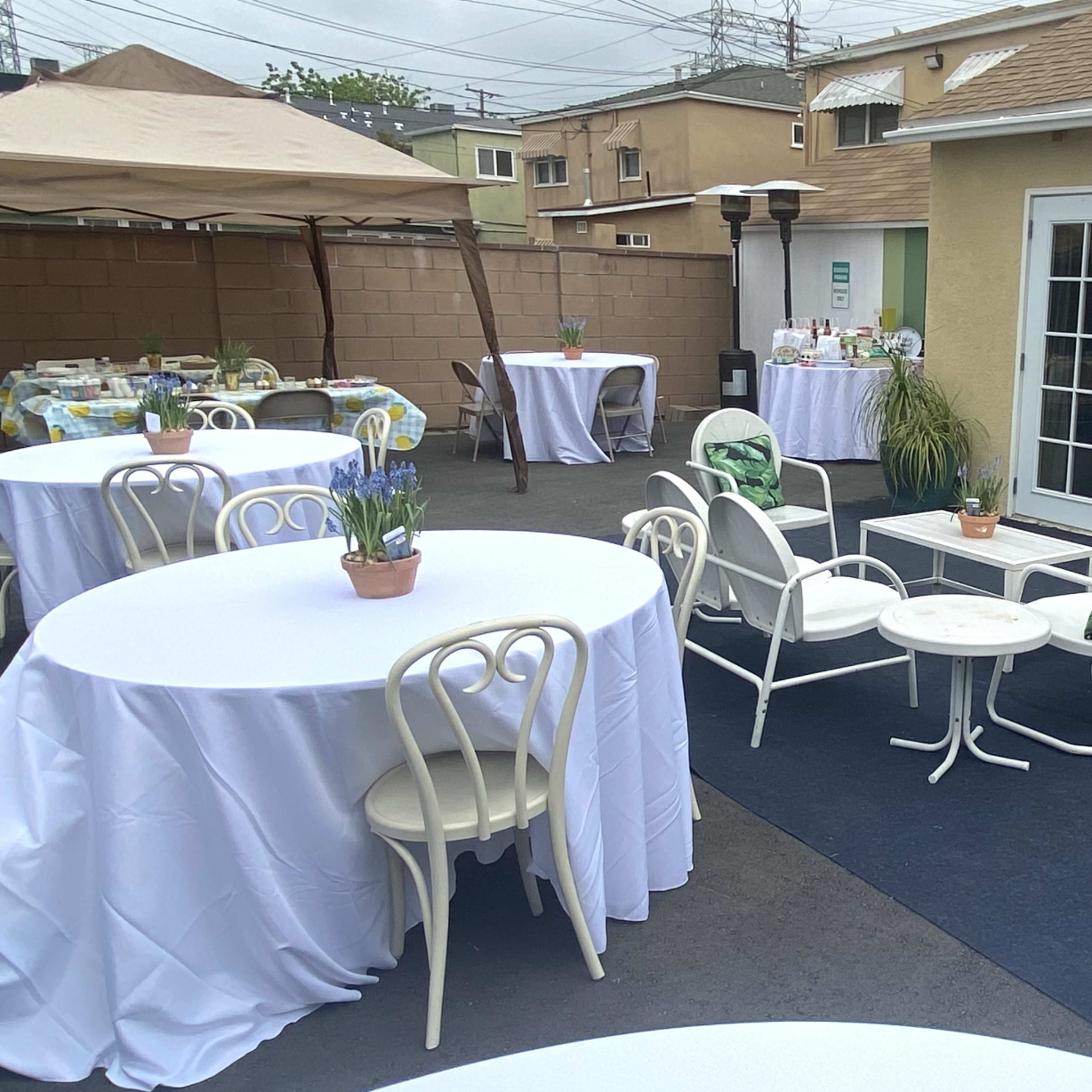An outdoor seating area features several round tables covered with white tablecloths, a tent, and two sets of double doors leading into a building.