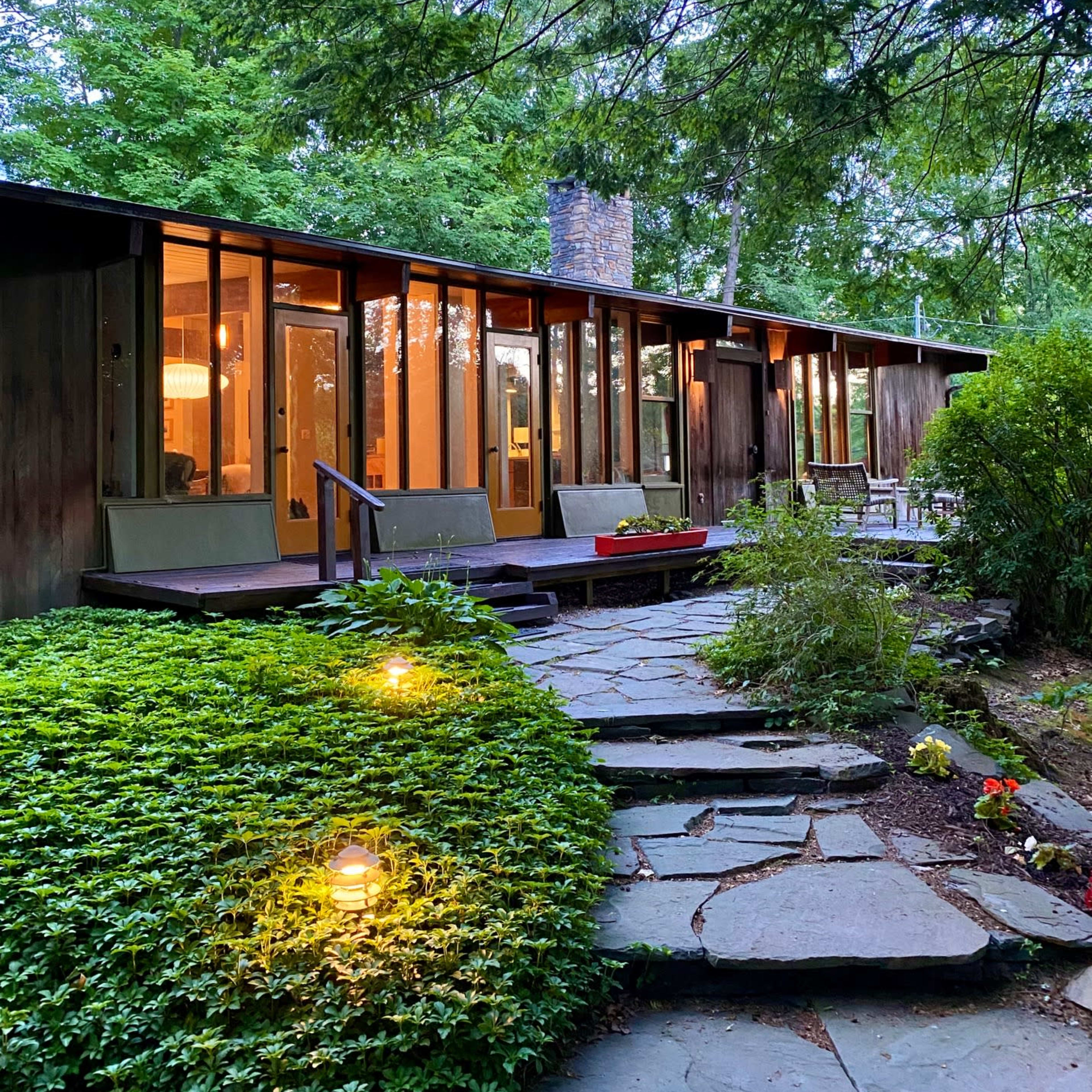 A stone pathway leads to a modern house with large windows, surrounded by greenery and trees.