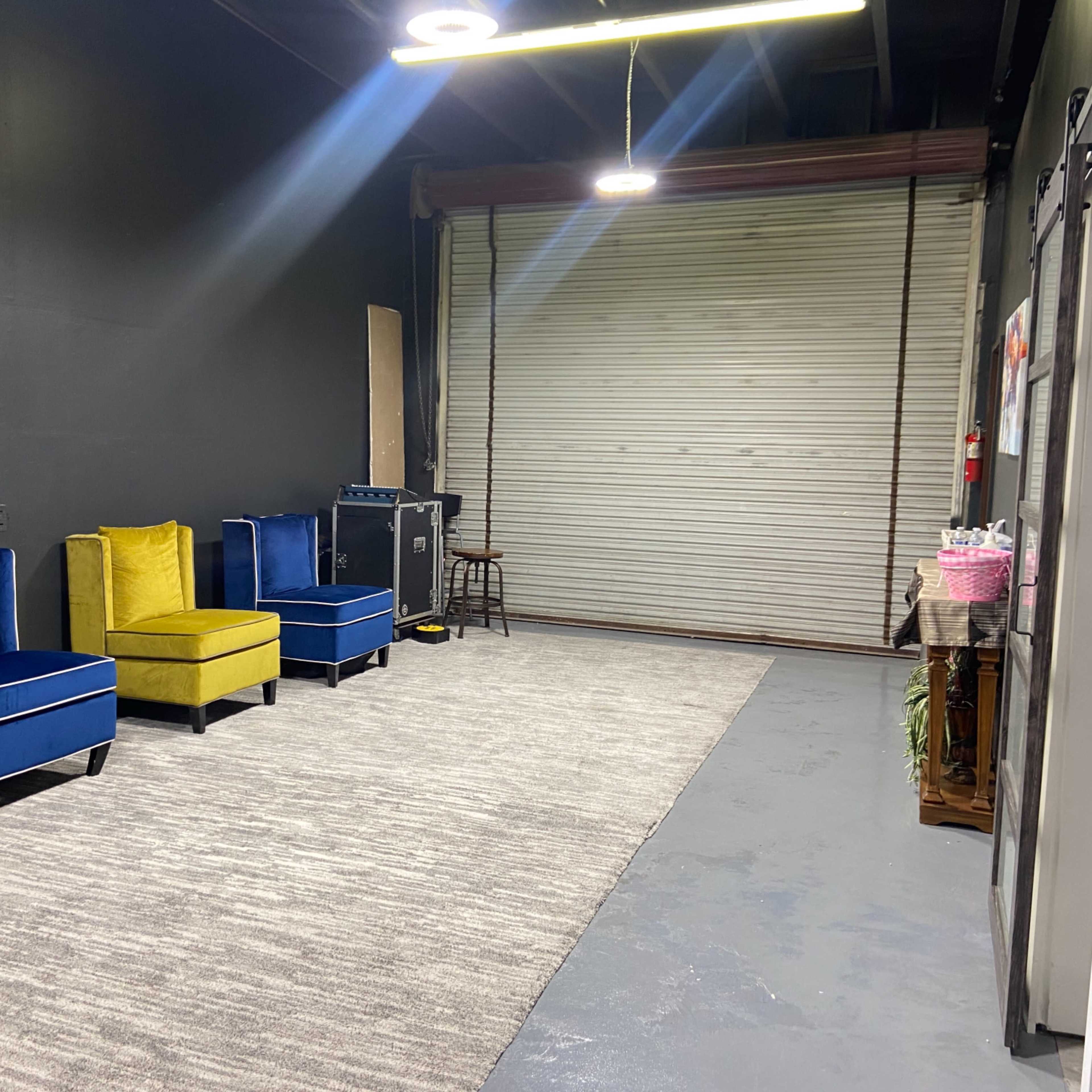 An interior space with two blue chairs and one yellow chair arranged along a gray rug, facing a closed metal garage door.