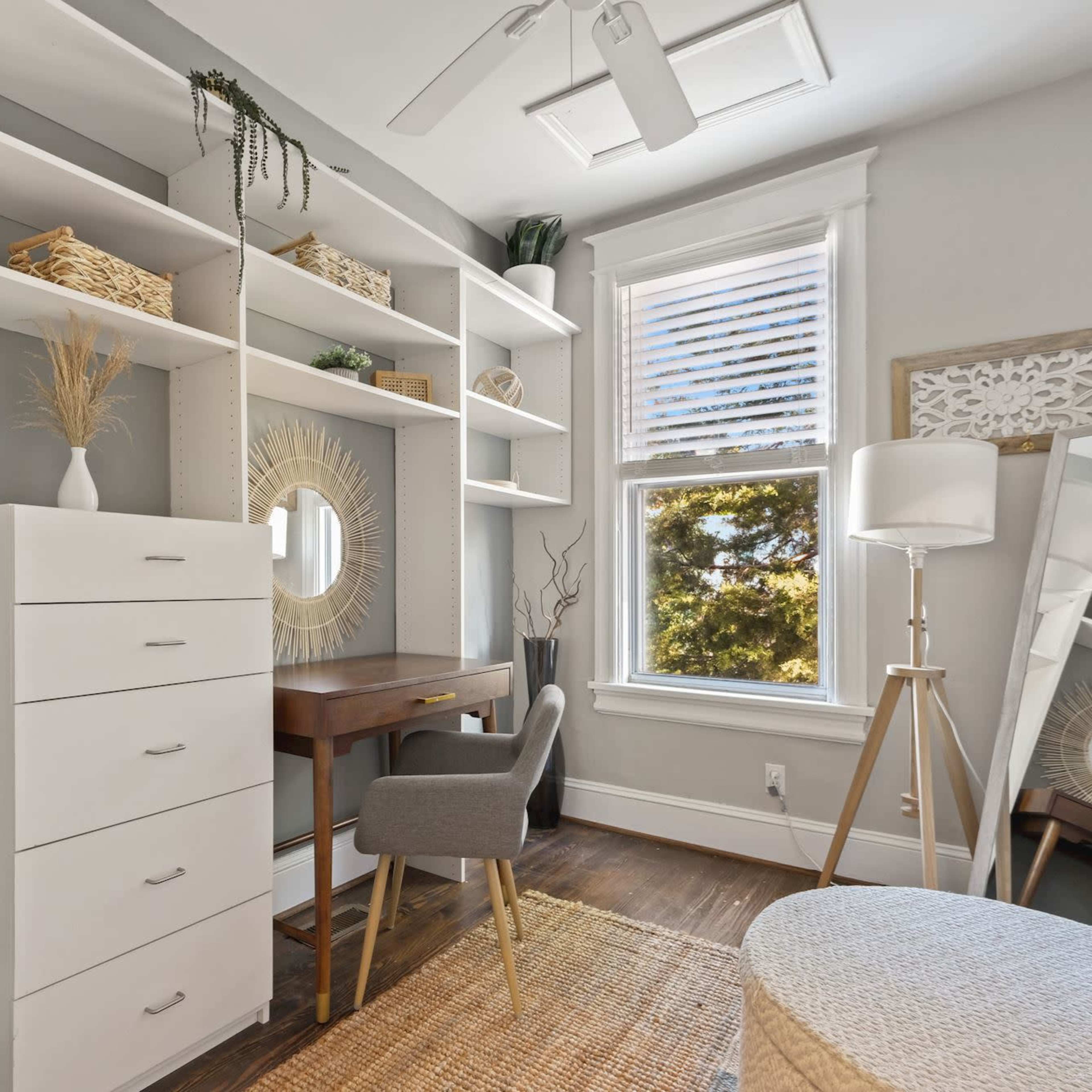 A modern home office with a desk, shelving unit, and a large mirror, illuminated by natural light from a window.