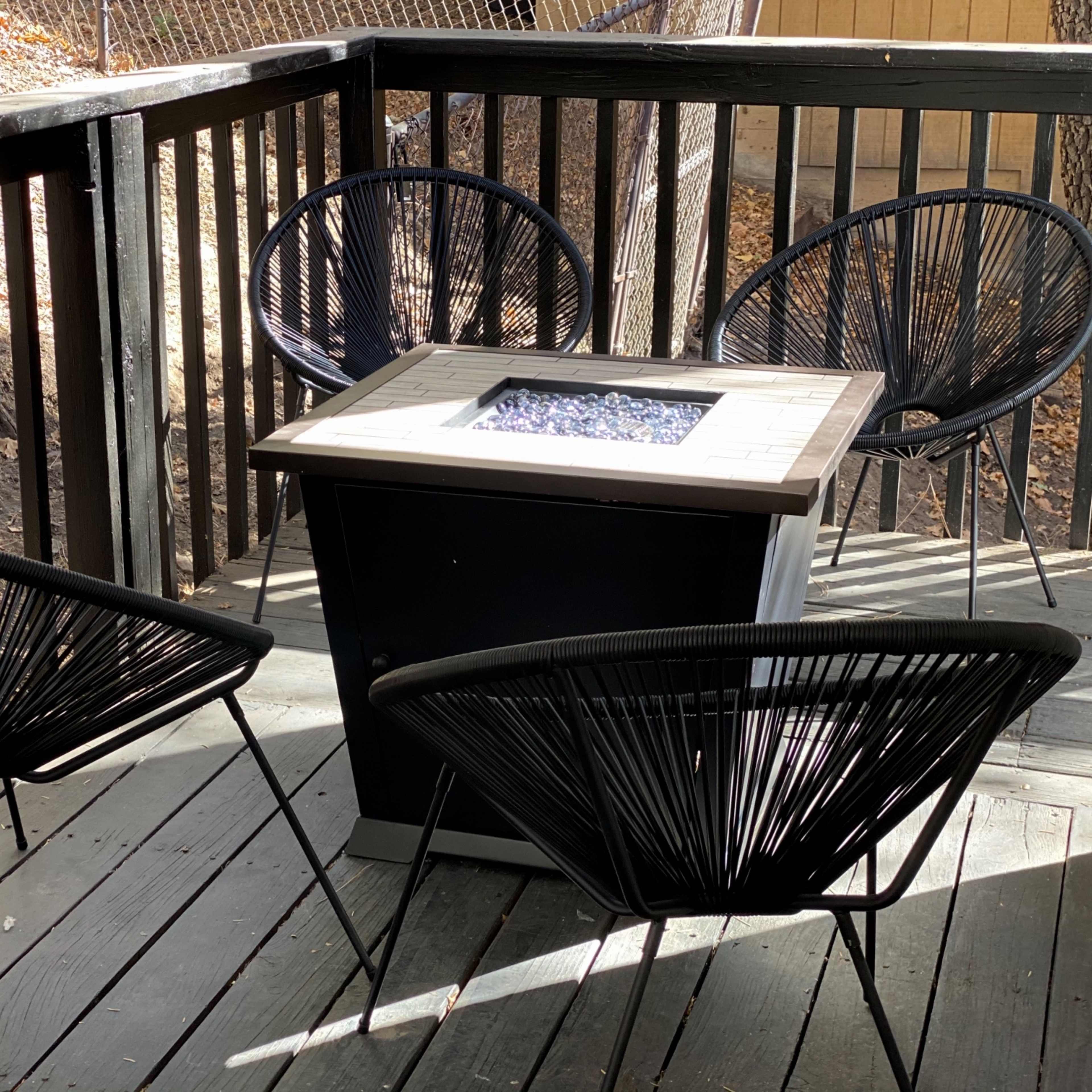 A patio area with four black wire chairs surrounding a square fire pit on a wooden deck.