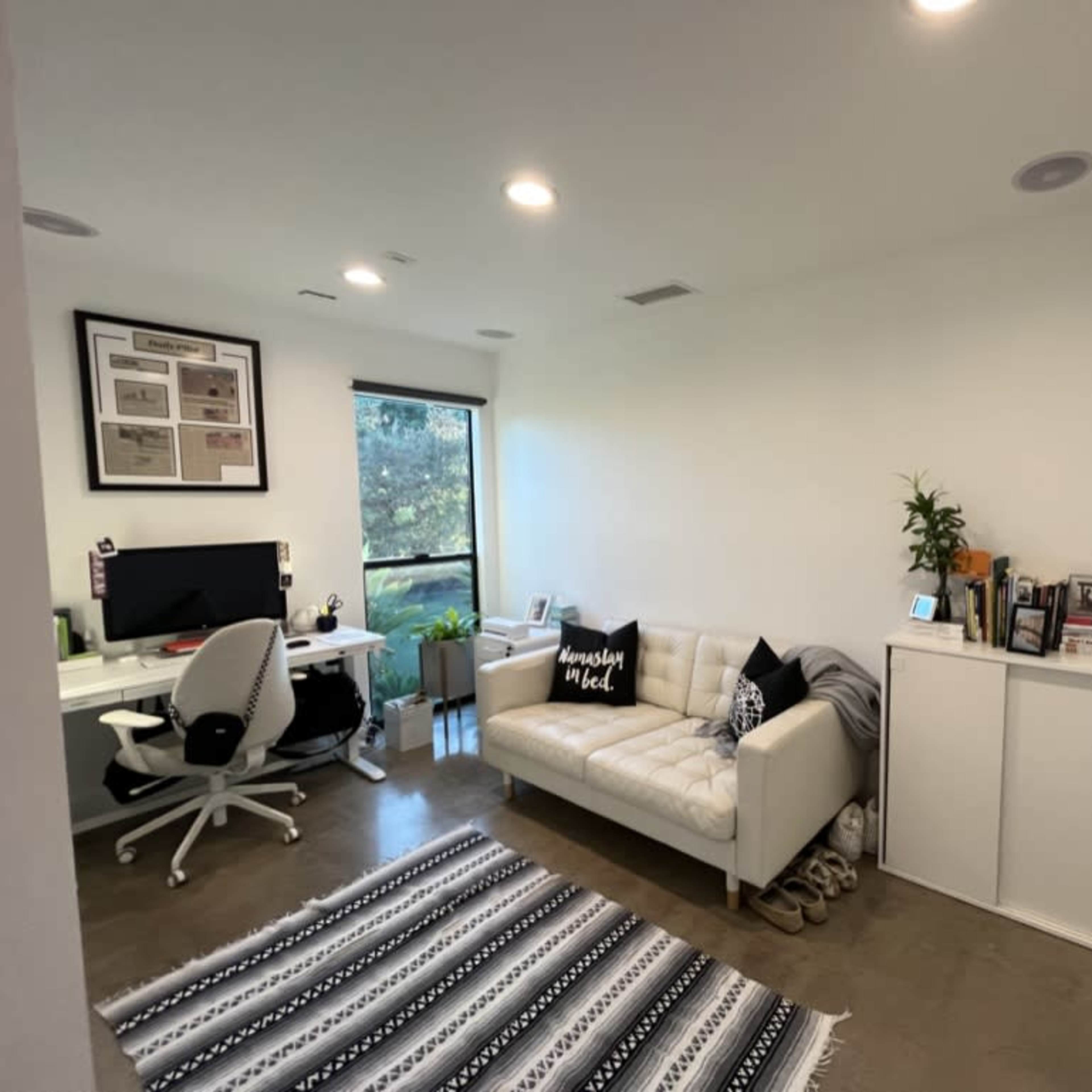 A modern office space featuring a white sofa, a black-and-white patterned rug, a desk with a computer, and a window allowing natural light.