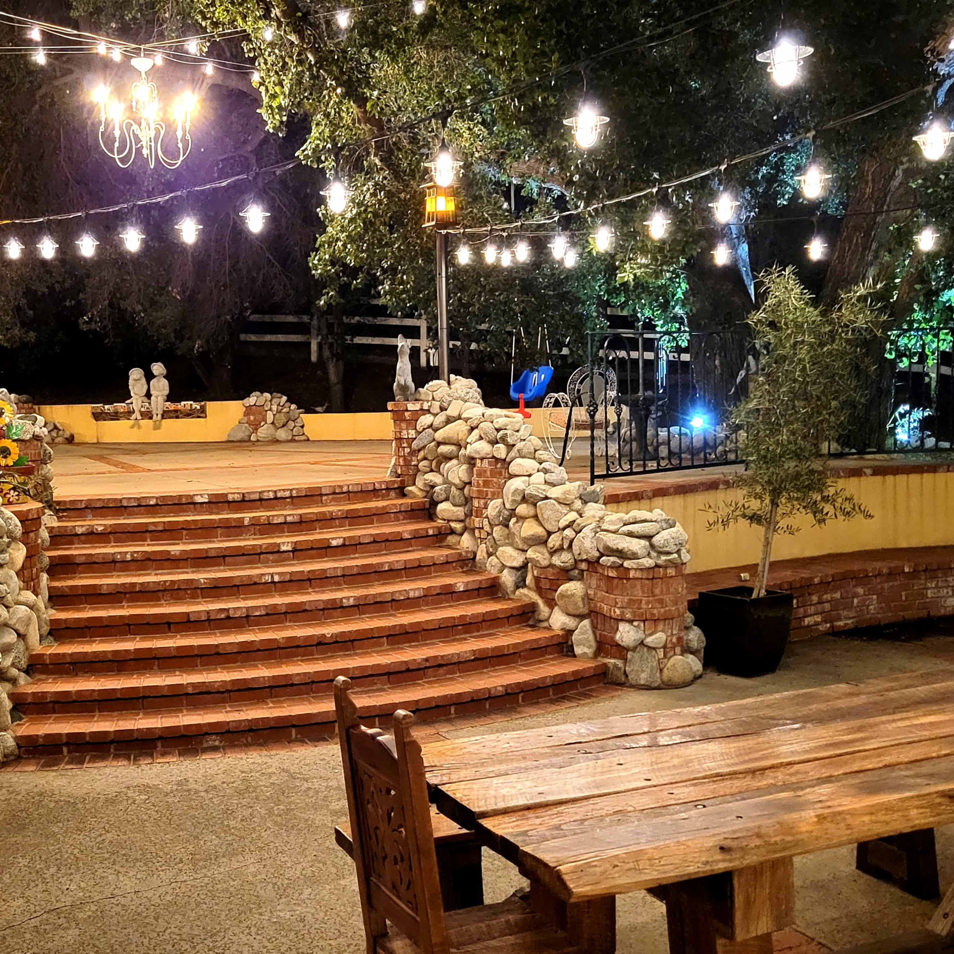 An outdoor garden area with stone steps leading to a raised platform, surrounded by string lights and illuminated by lanterns.