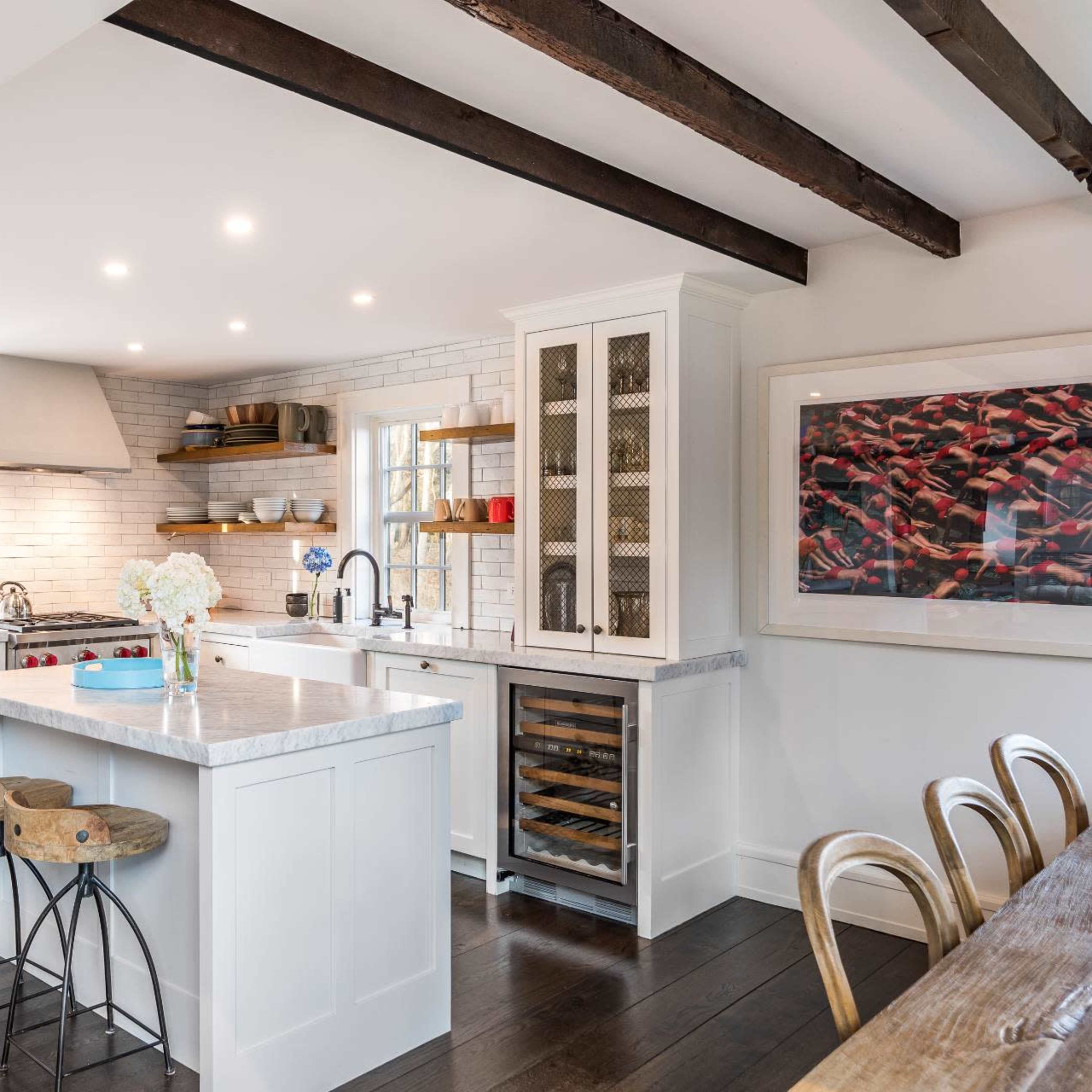 A modern kitchen features a central island, white cabinetry, stainless steel appliances, and exposed wooden beams.