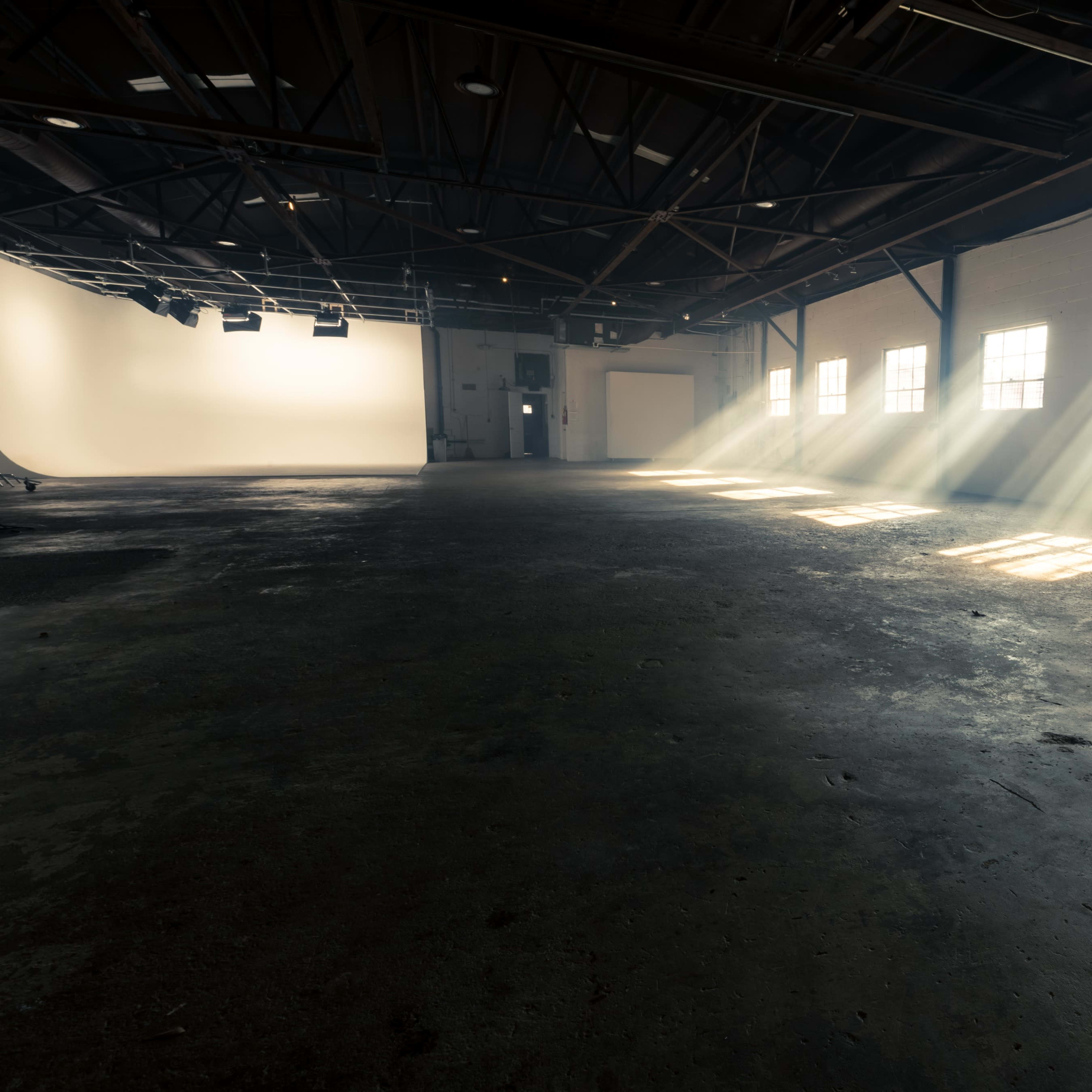 A spacious, empty warehouse with sunlight streaming through the windows, casting rays onto the concrete floor.