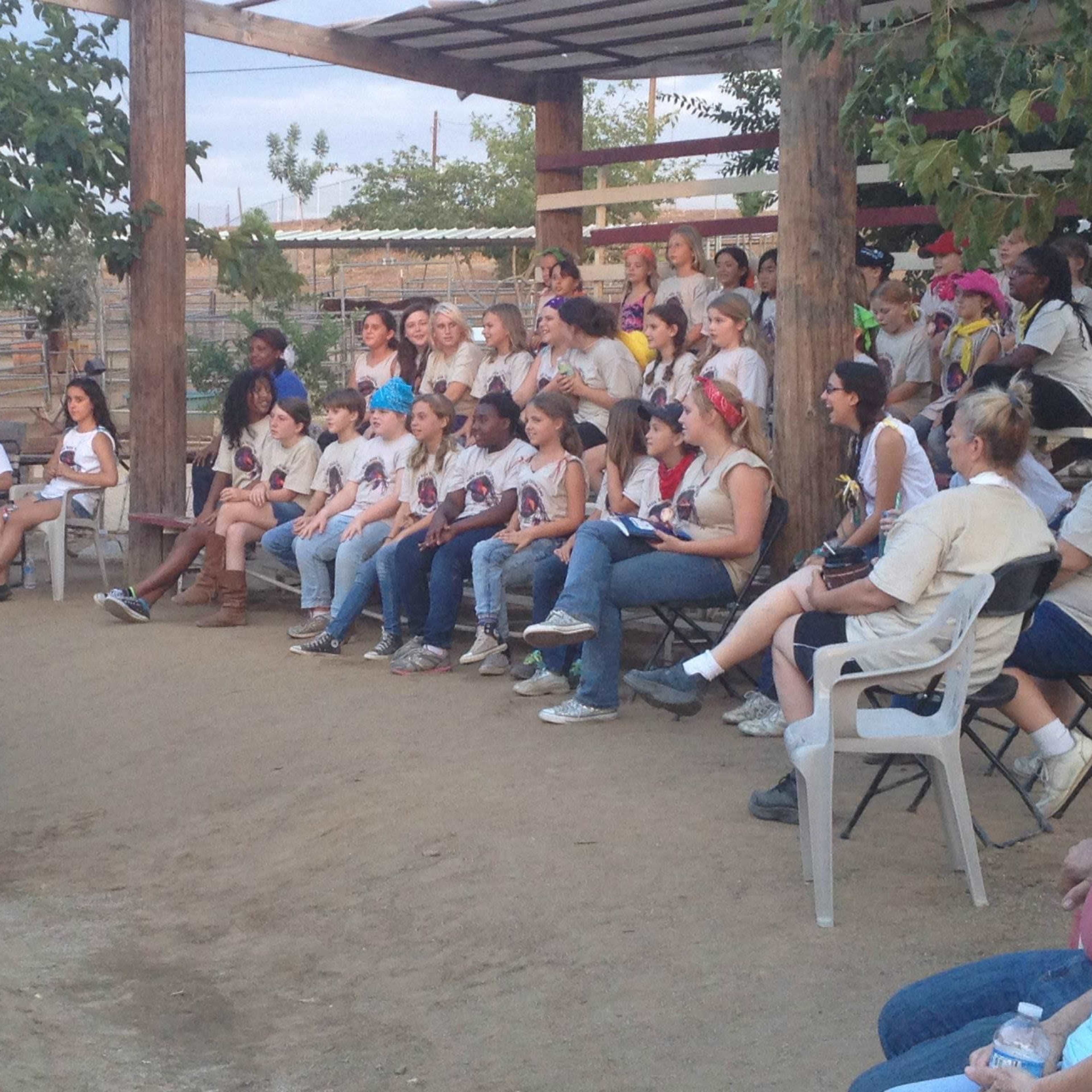 A group of children and adults sit in a semi-circle on chairs, attentively listening to a person standing in front of them in an outdoor setting.