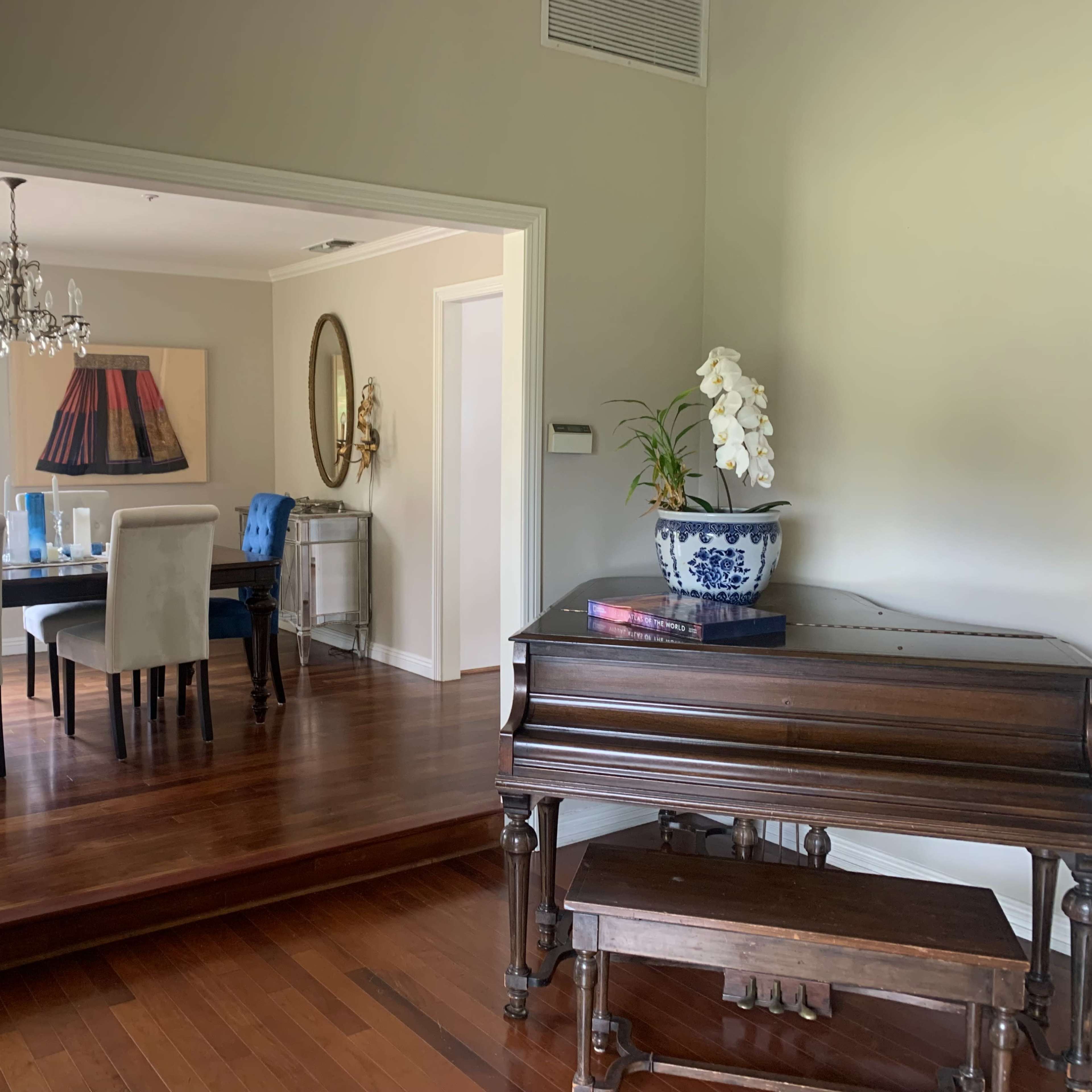 A wooden piano stands in the foreground while a dining area with a table and chairs is visible through an open doorway.