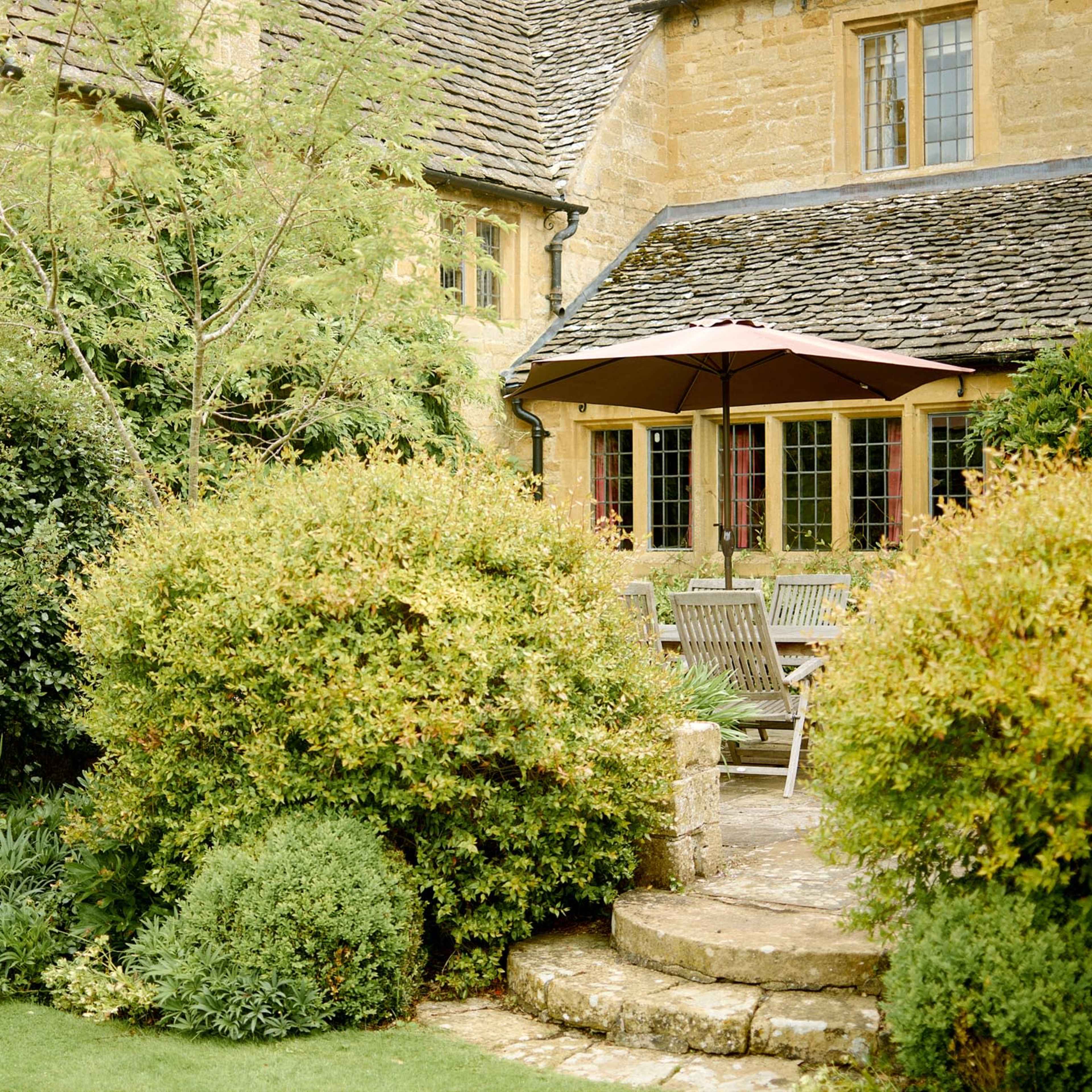 A stone house with a patio and an umbrella is surrounded by neatly trimmed bushes and greenery.