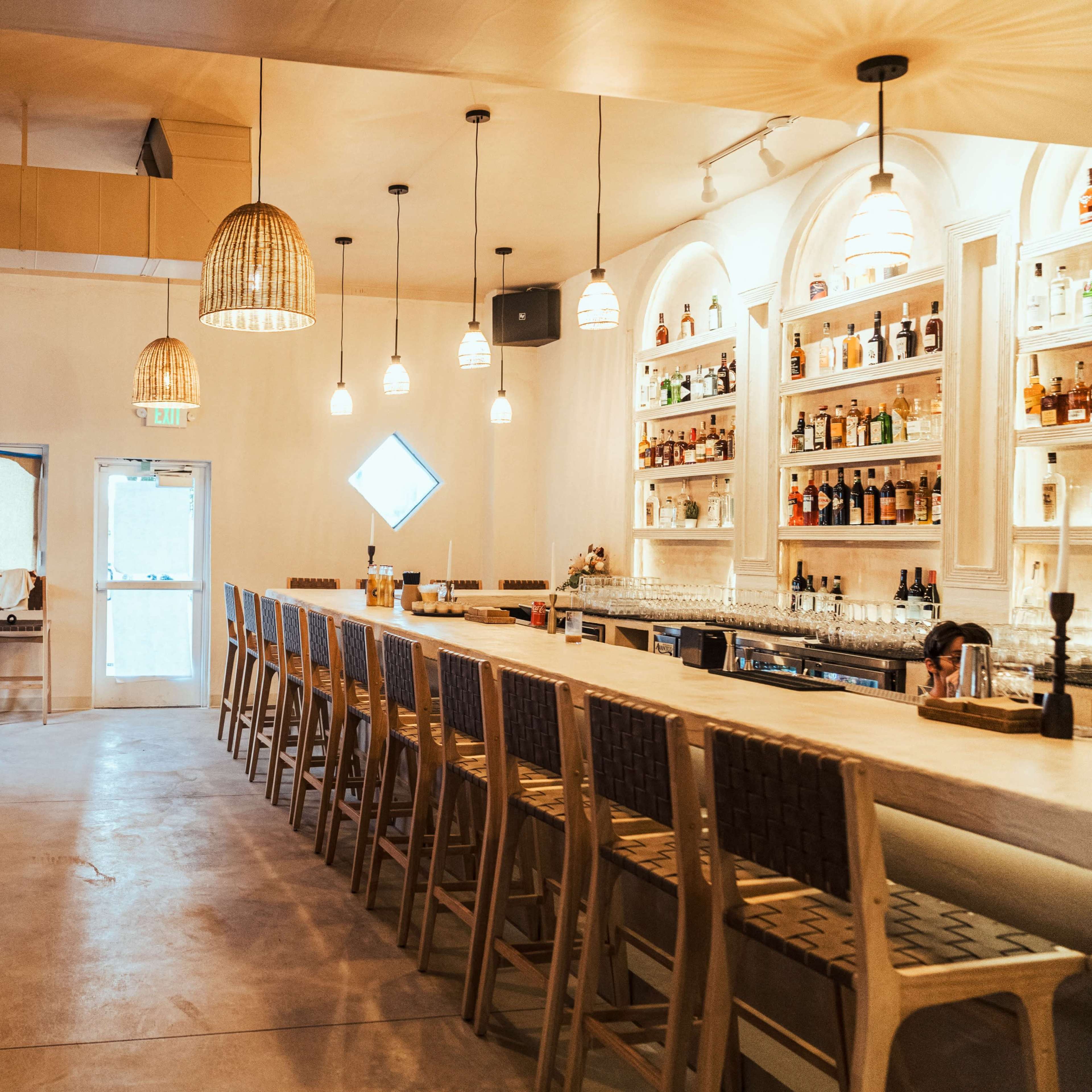 A spacious bar area with a long wooden counter, overhead lighting fixtures, and a wall of shelves stocked with various bottles.