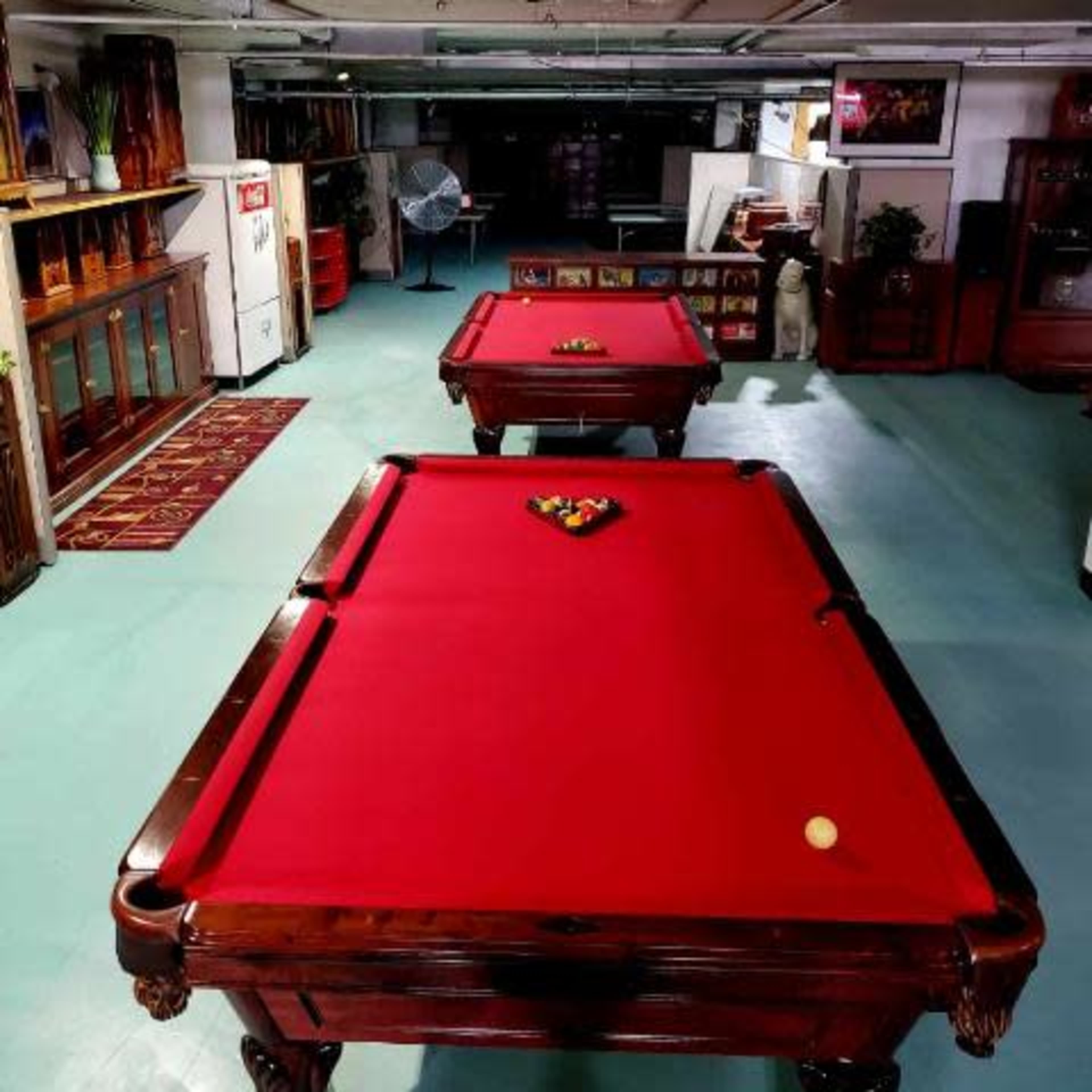 Two pool tables with red felt set up in a spacious room featuring wooden furniture and a fan.