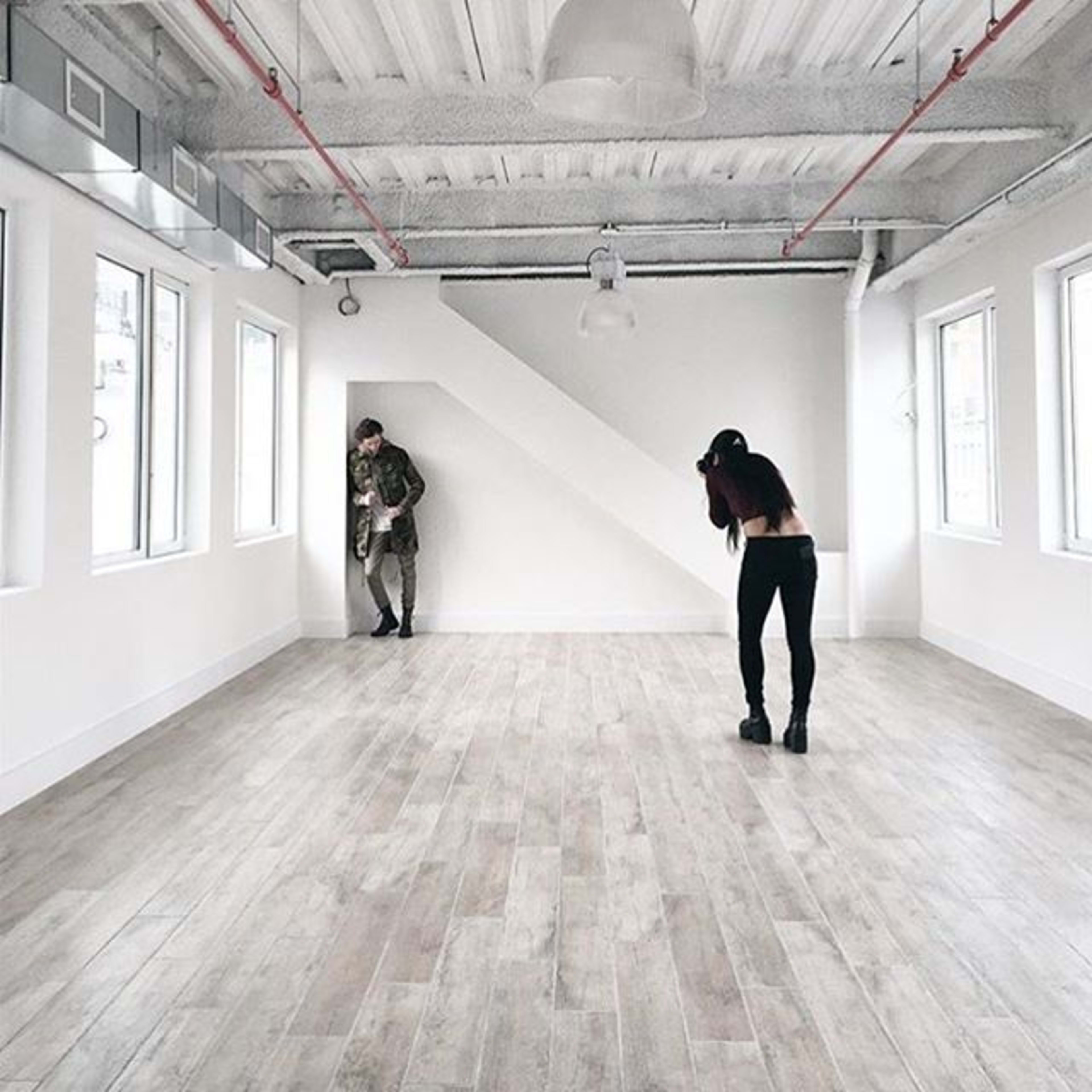 A woman takes photos of a man posing in a spacious, minimally furnished room with large windows and wooden flooring.