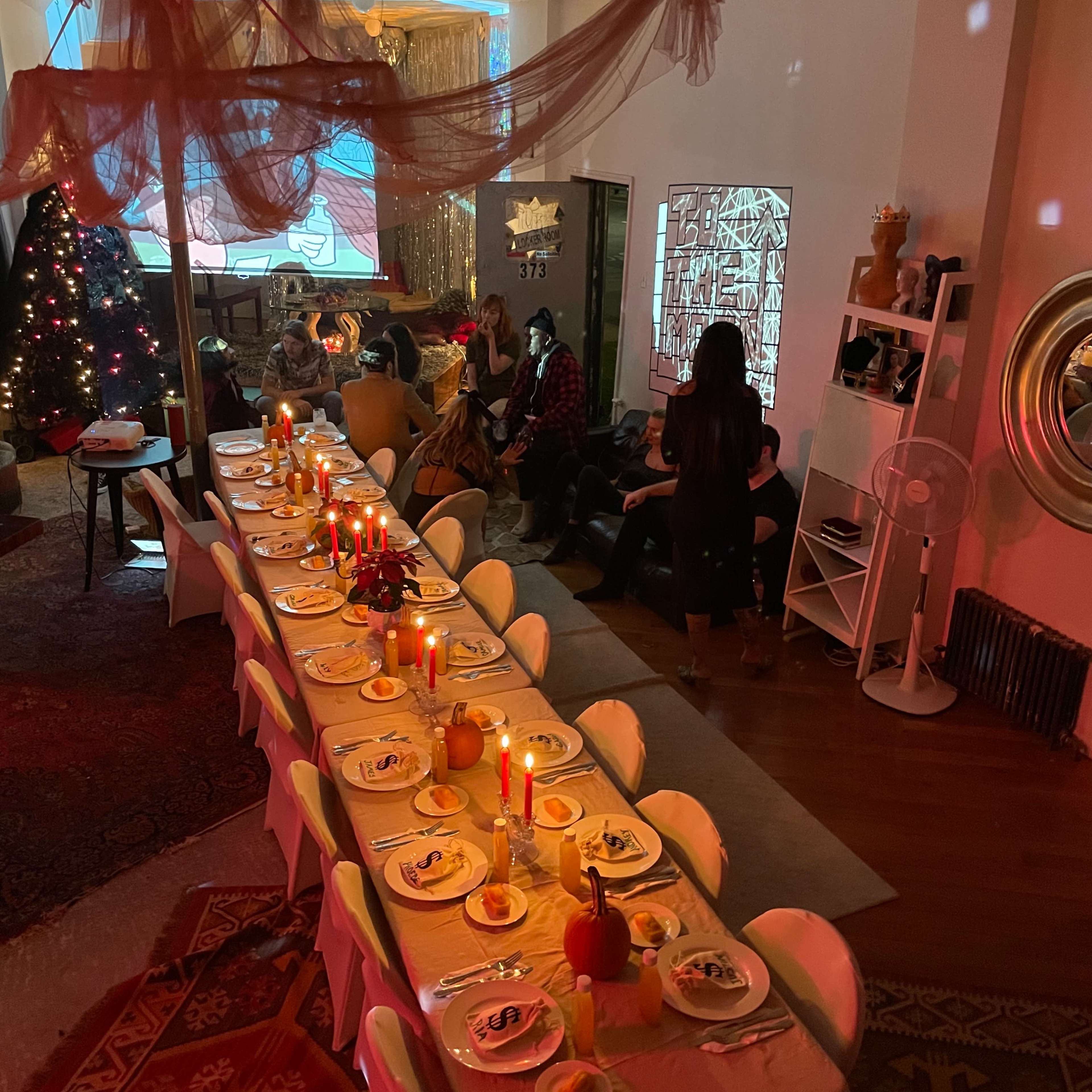 A long dining table is set with candles and plates, while guests gather in a decorated room with a Christmas tree and festive decorations.