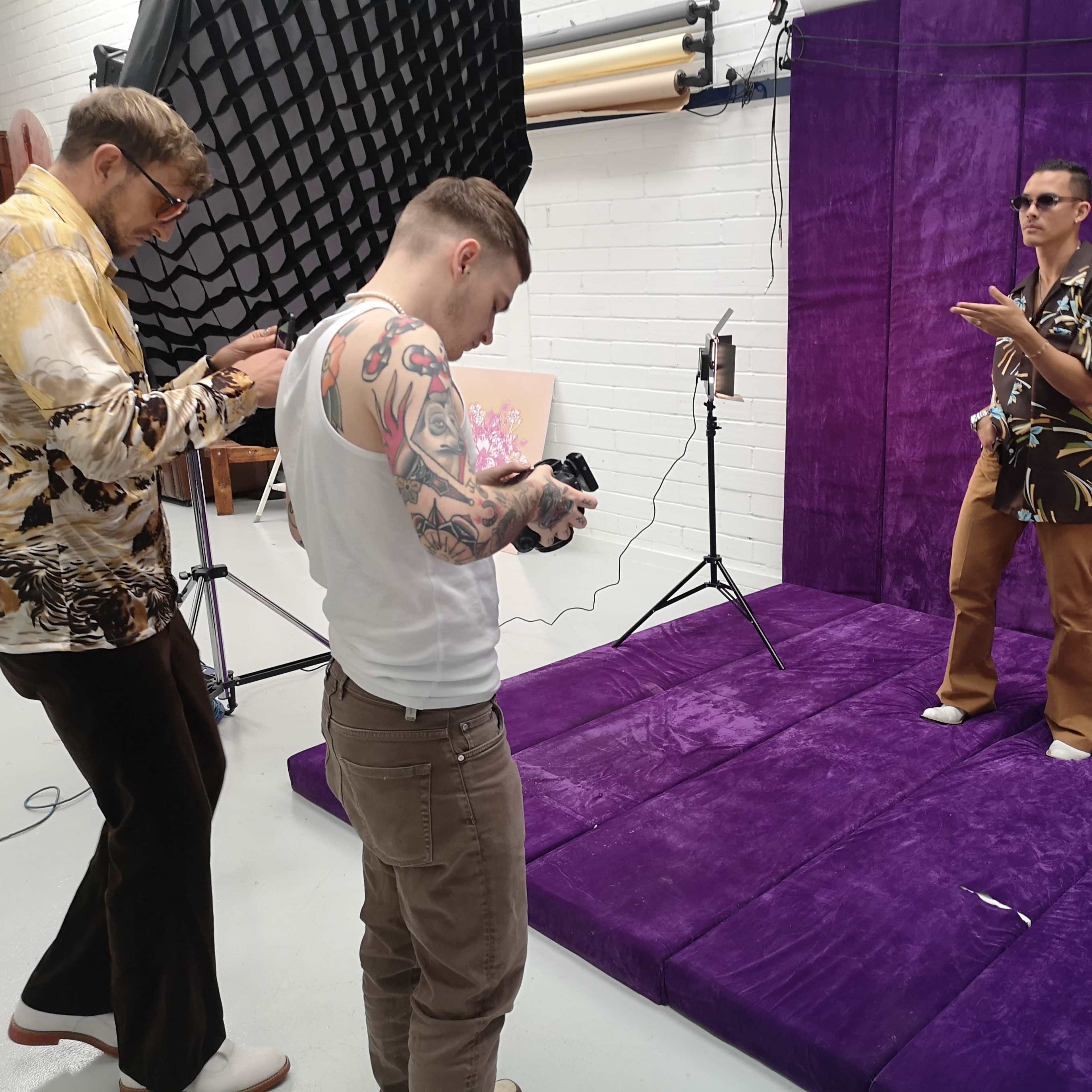 Three individuals are in a studio setting, one posing in front of a purple backdrop while the others are taking photos and adjusting their cameras.