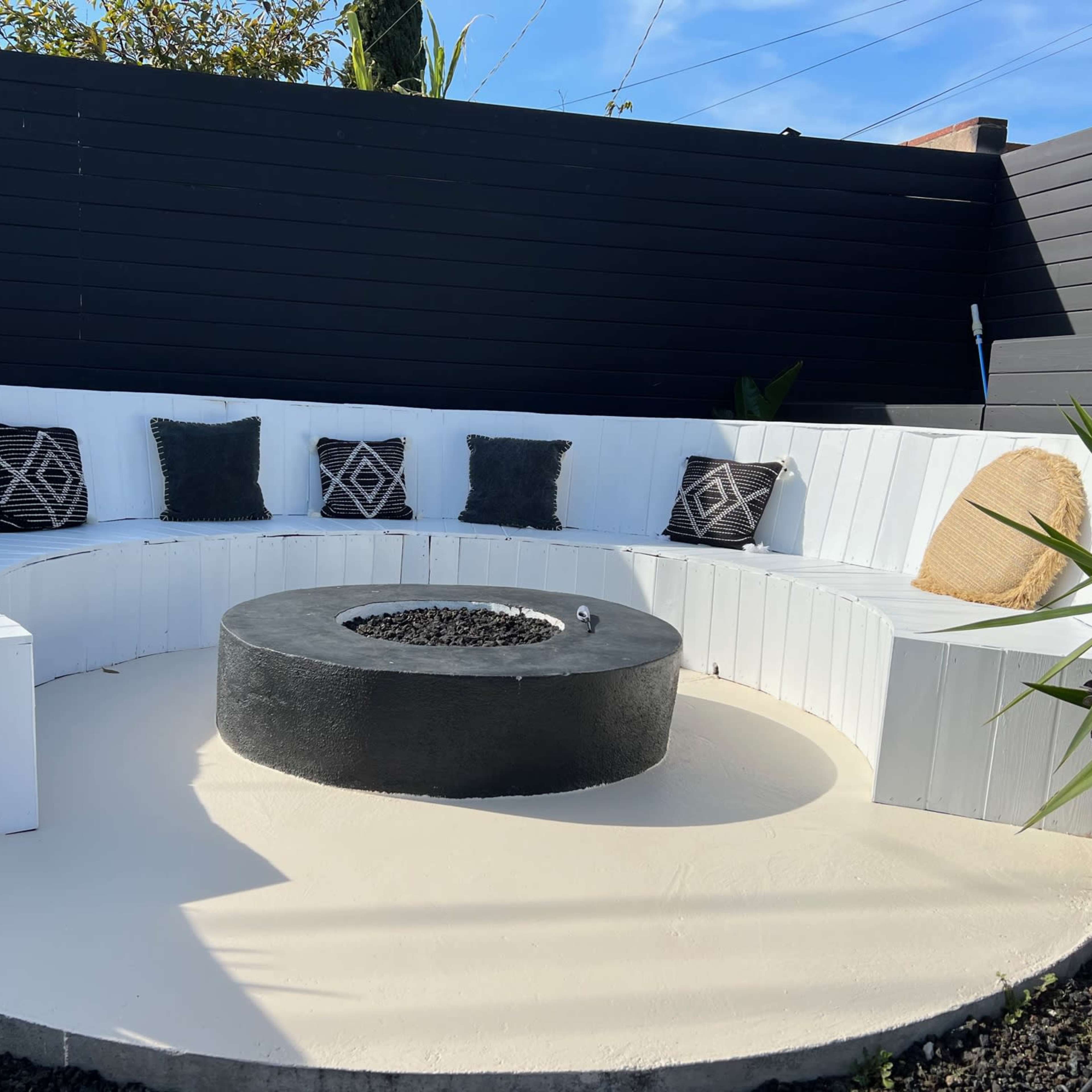 A circular outdoor seating area with white benches, black cushions, and a central fire pit surrounded by concrete.