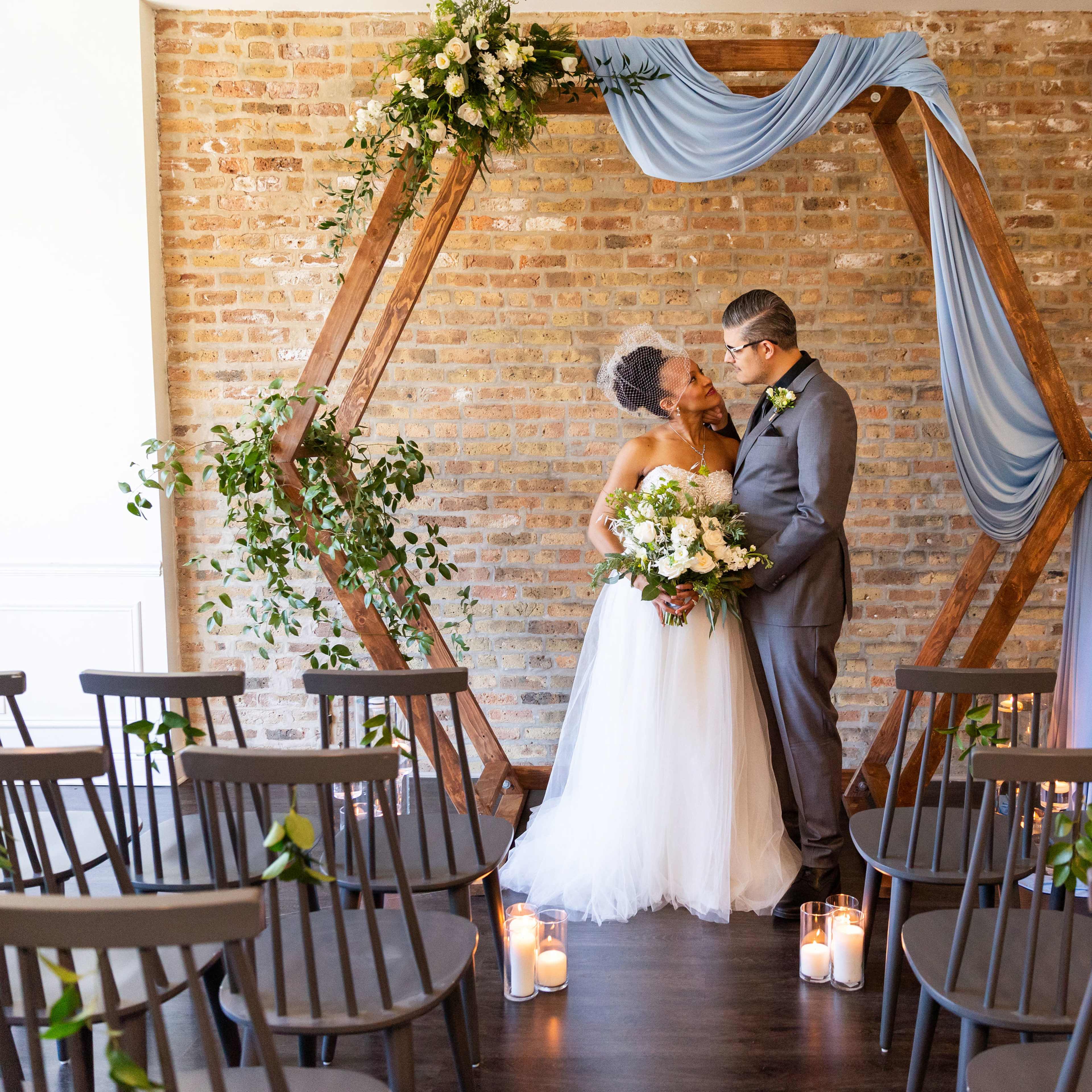 A bride and groom kiss under a floral arch in a brick-walled venue with chairs arranged for a wedding ceremony.