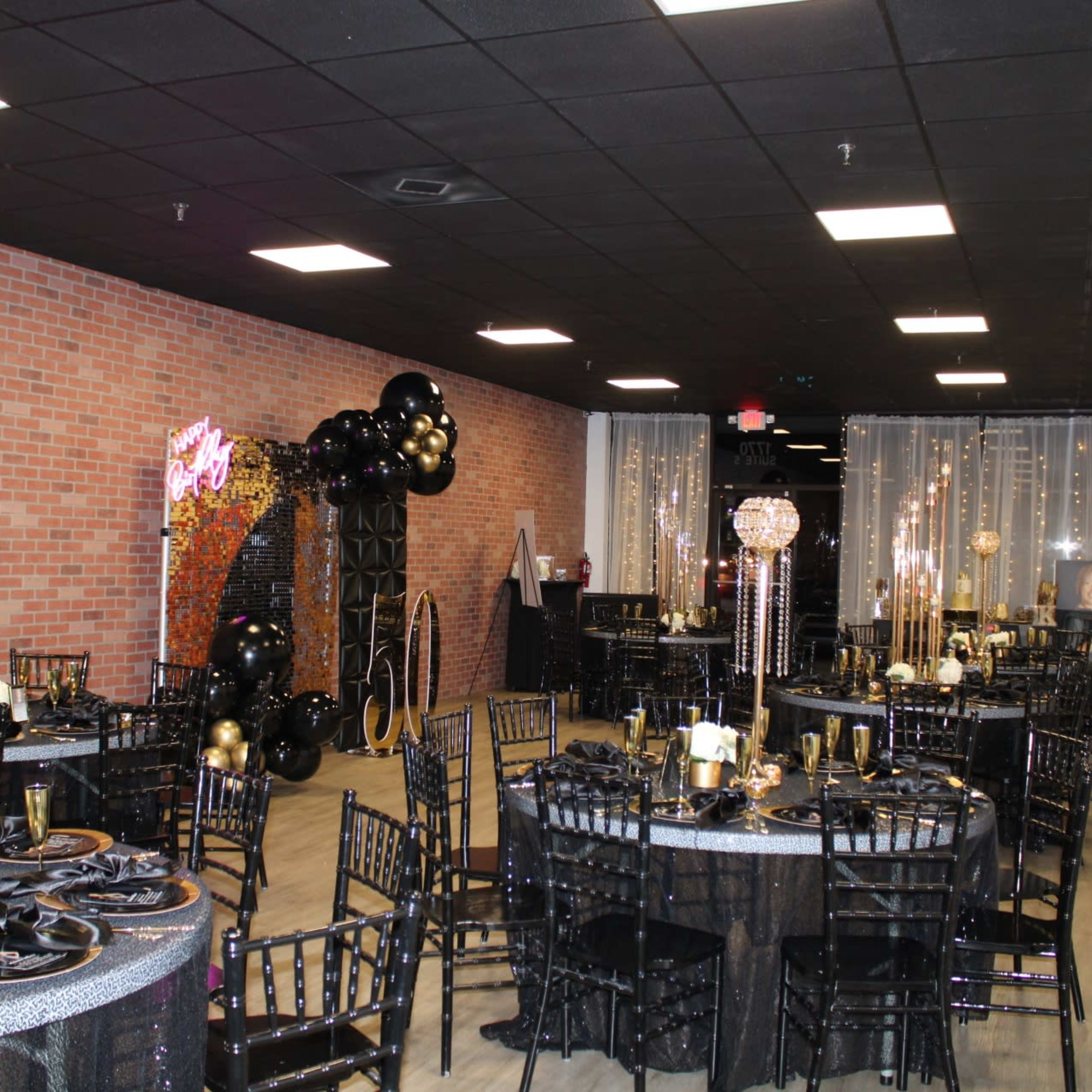 A decorated event space featuring black and gold themed tables, chairs, and decor, with a backdrop of balloons and lights.