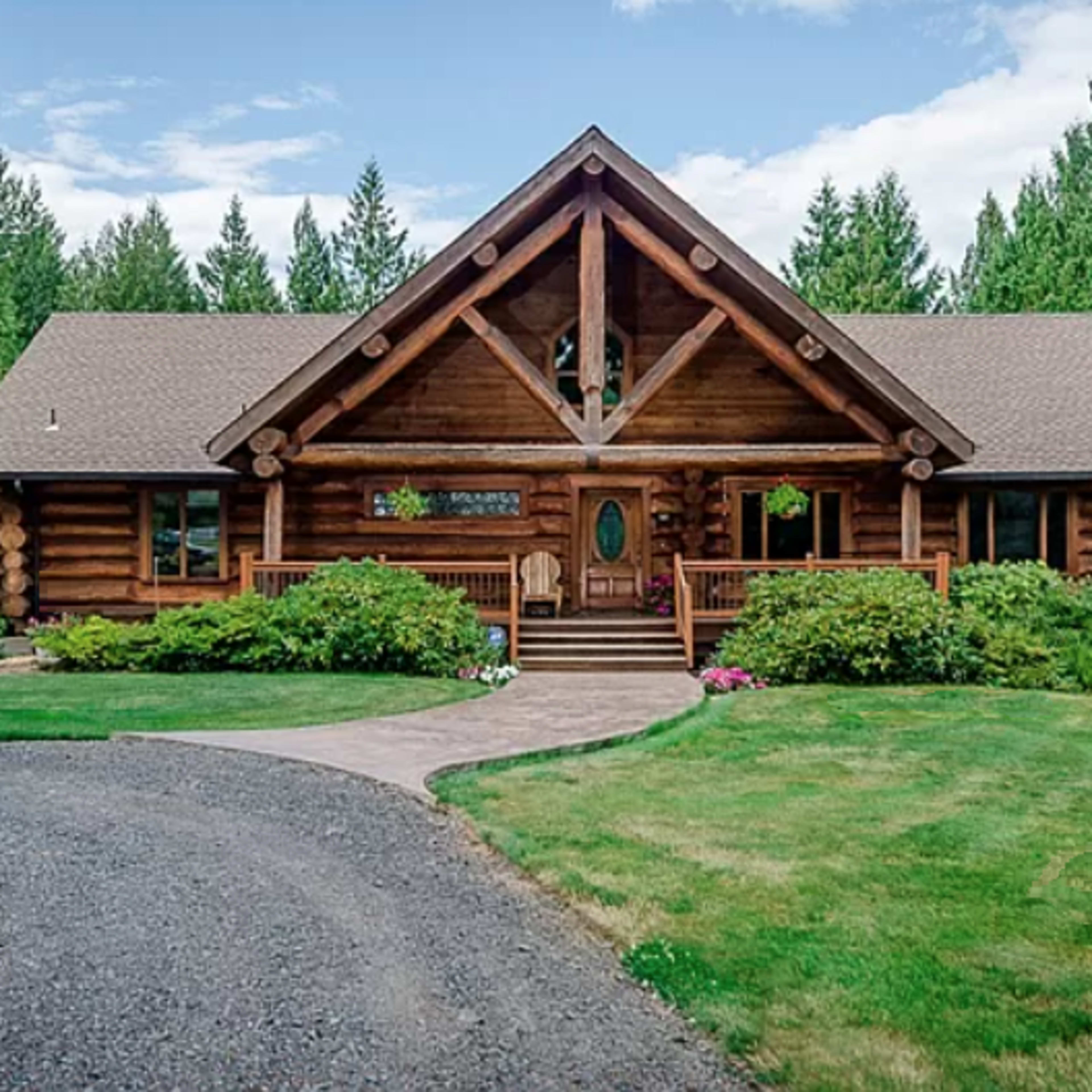 A log cabin-style house sits amid green trees and landscaped gardens, featuring a front porch and a stone walkway.