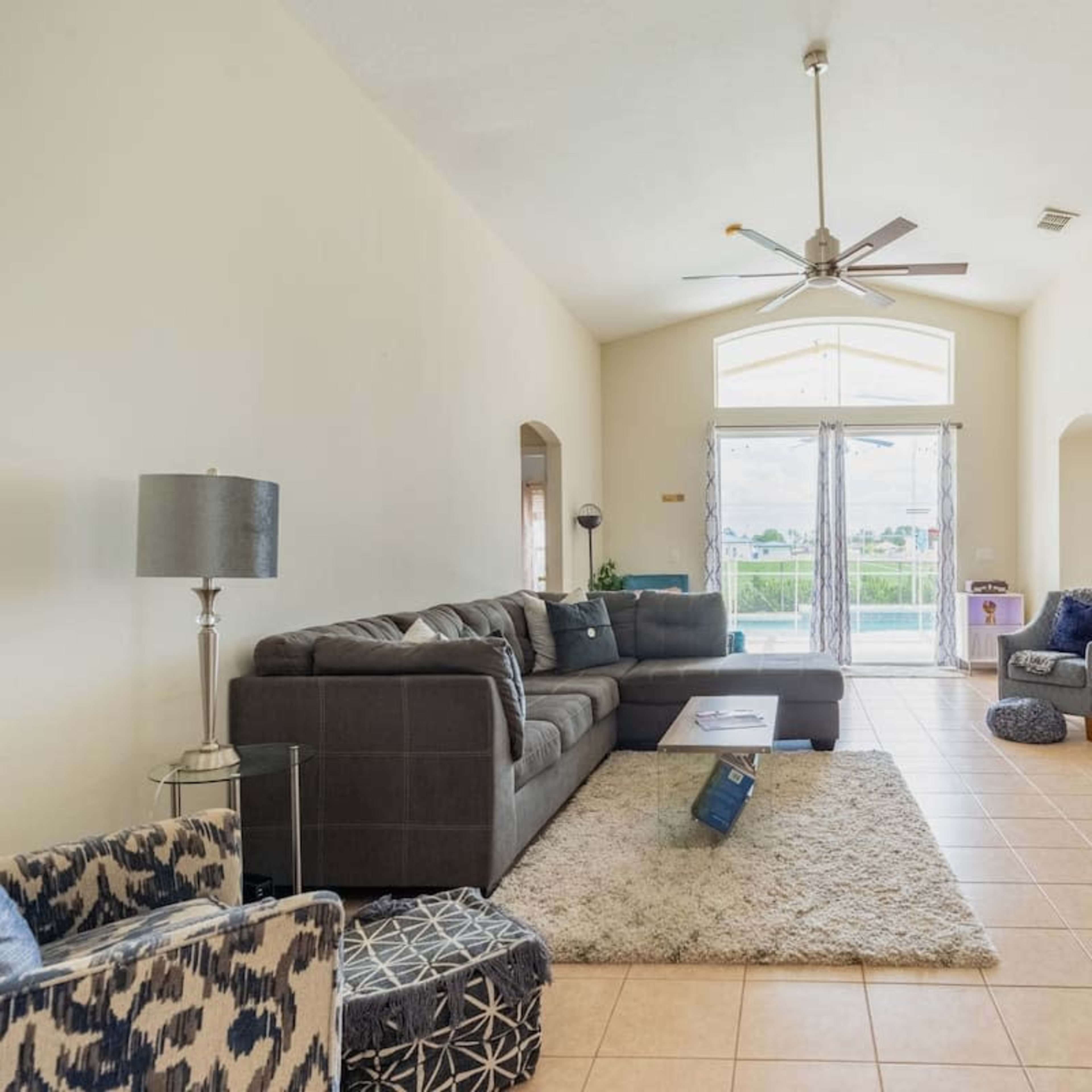 A living room with a large sectional couch, a coffee table, and a television, featuring tile flooring and large windows that allow natural light.