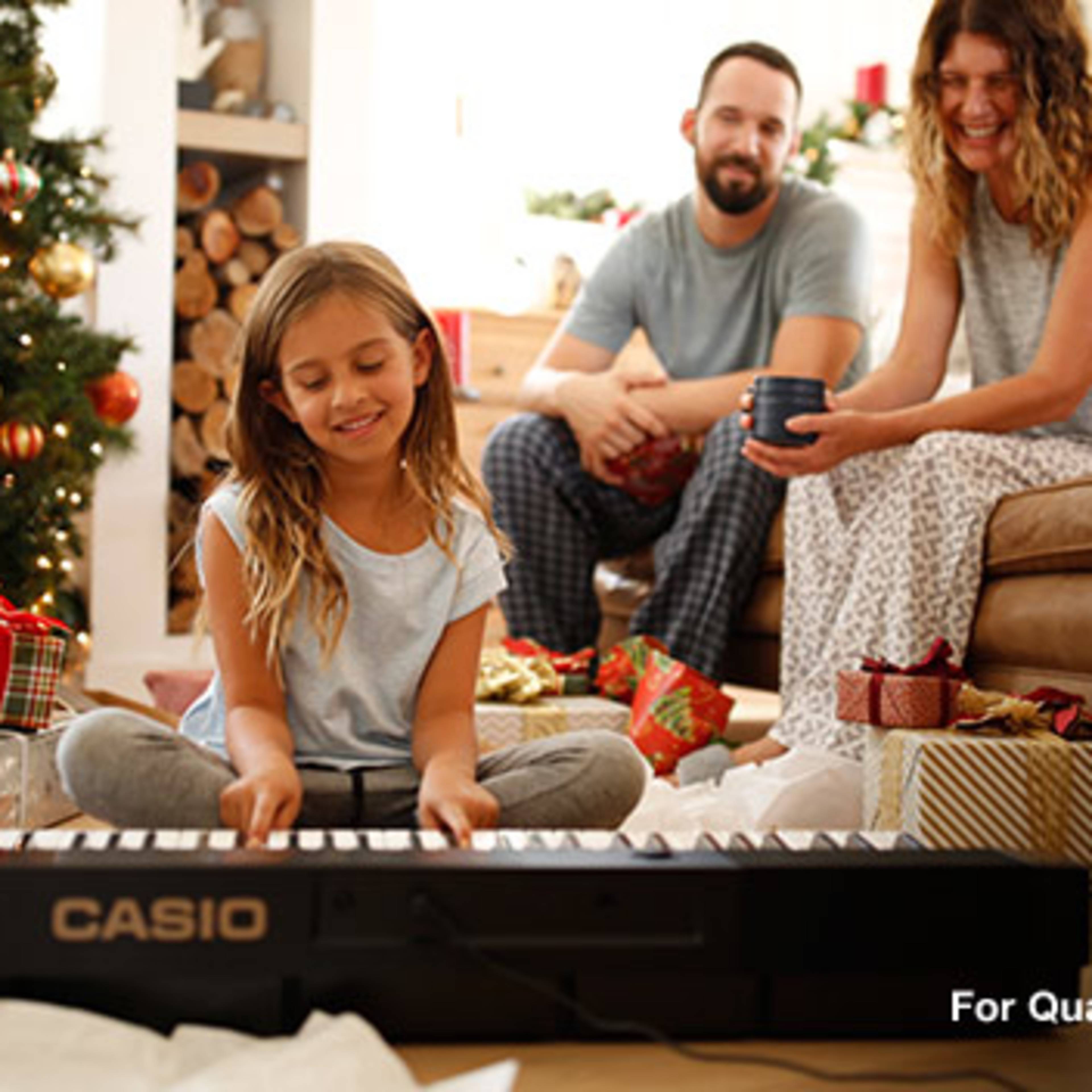 A young girl plays a piano while her parents sit on the couch surrounded by holiday decorations and gifts.