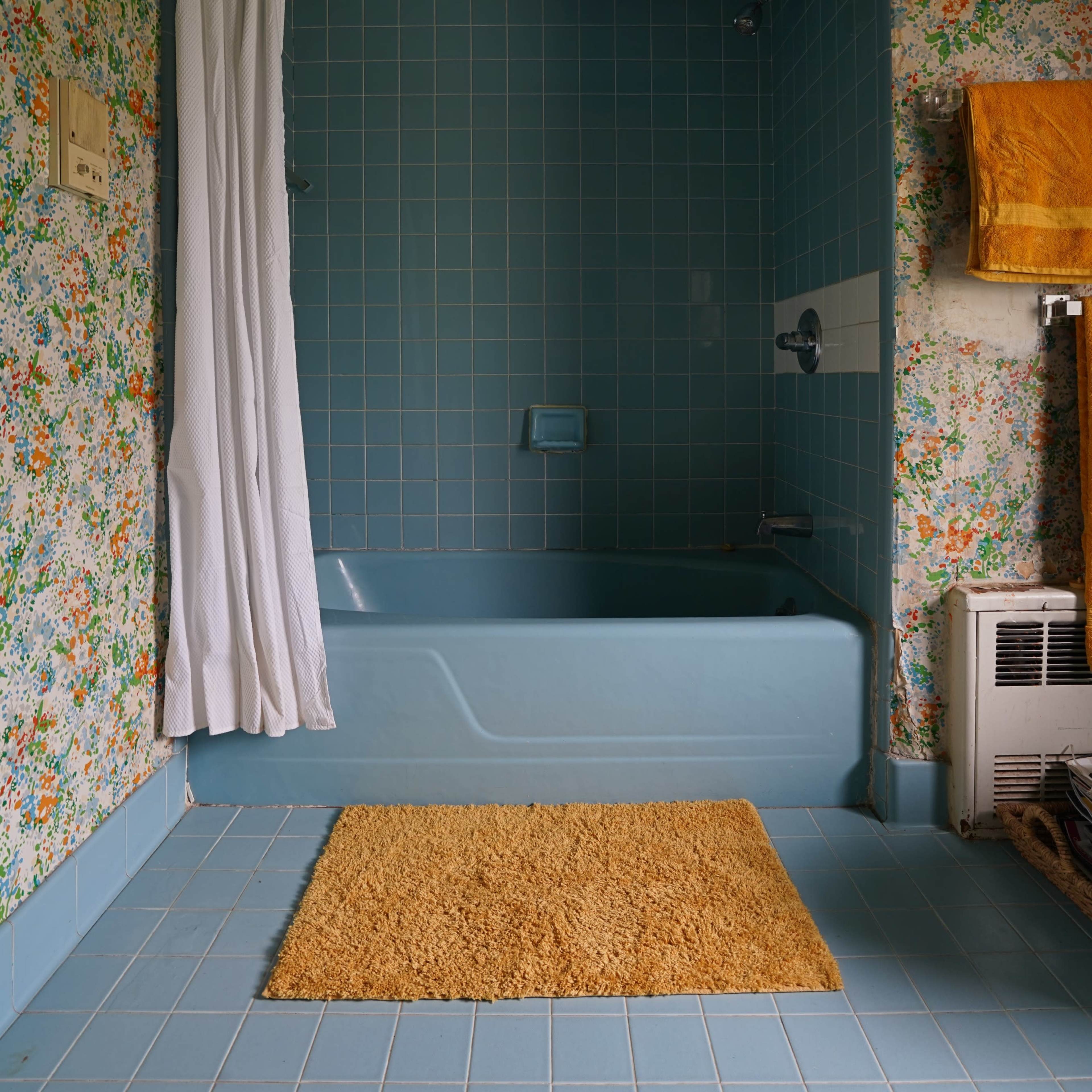 A bathroom featuring a blue bathtub, floral wallpaper, and a beige bath mat on a blue tiled floor.