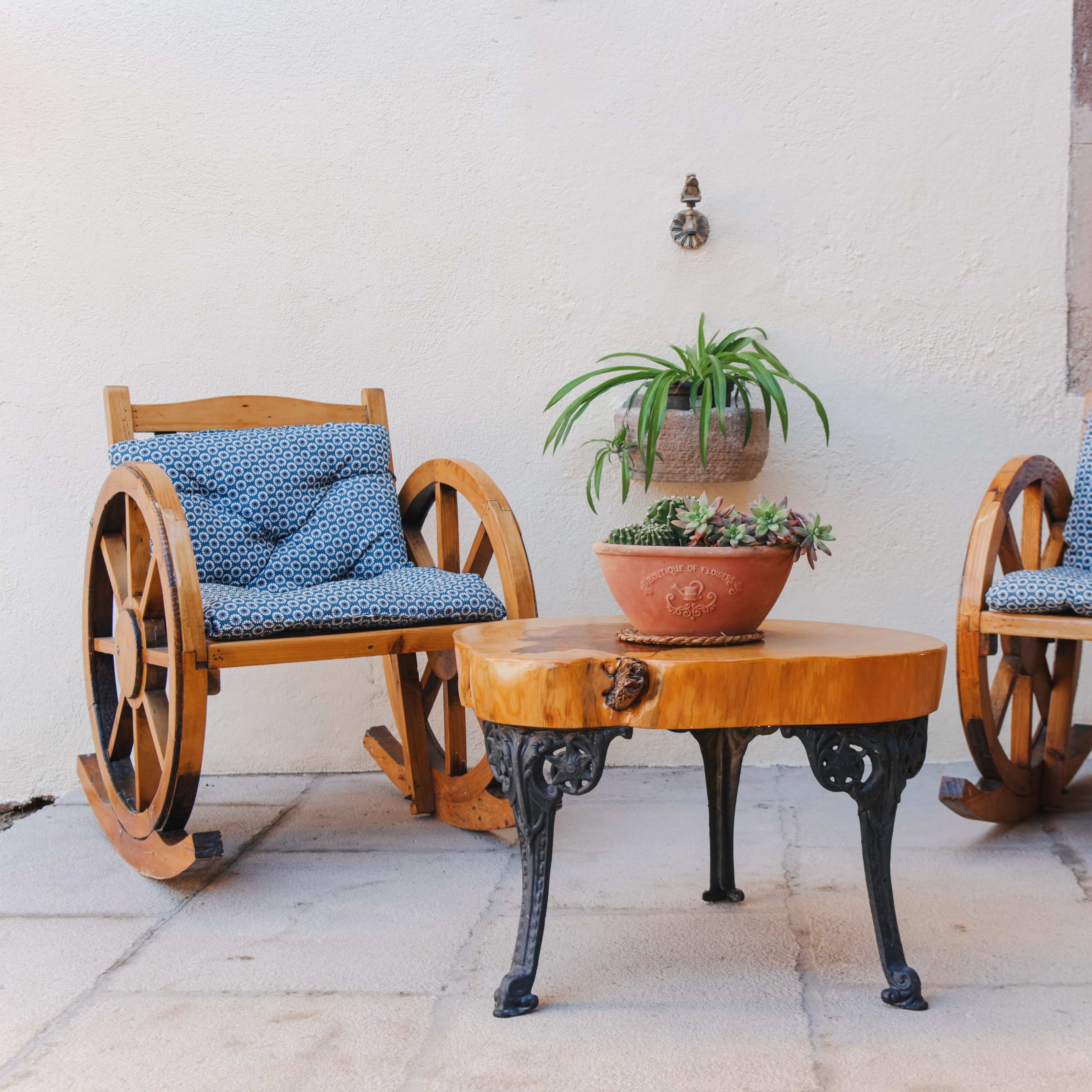 Two wooden rocking chairs with blue cushioned seats flanking a round wooden table with a potted plant on top, set against a white wall with a window.